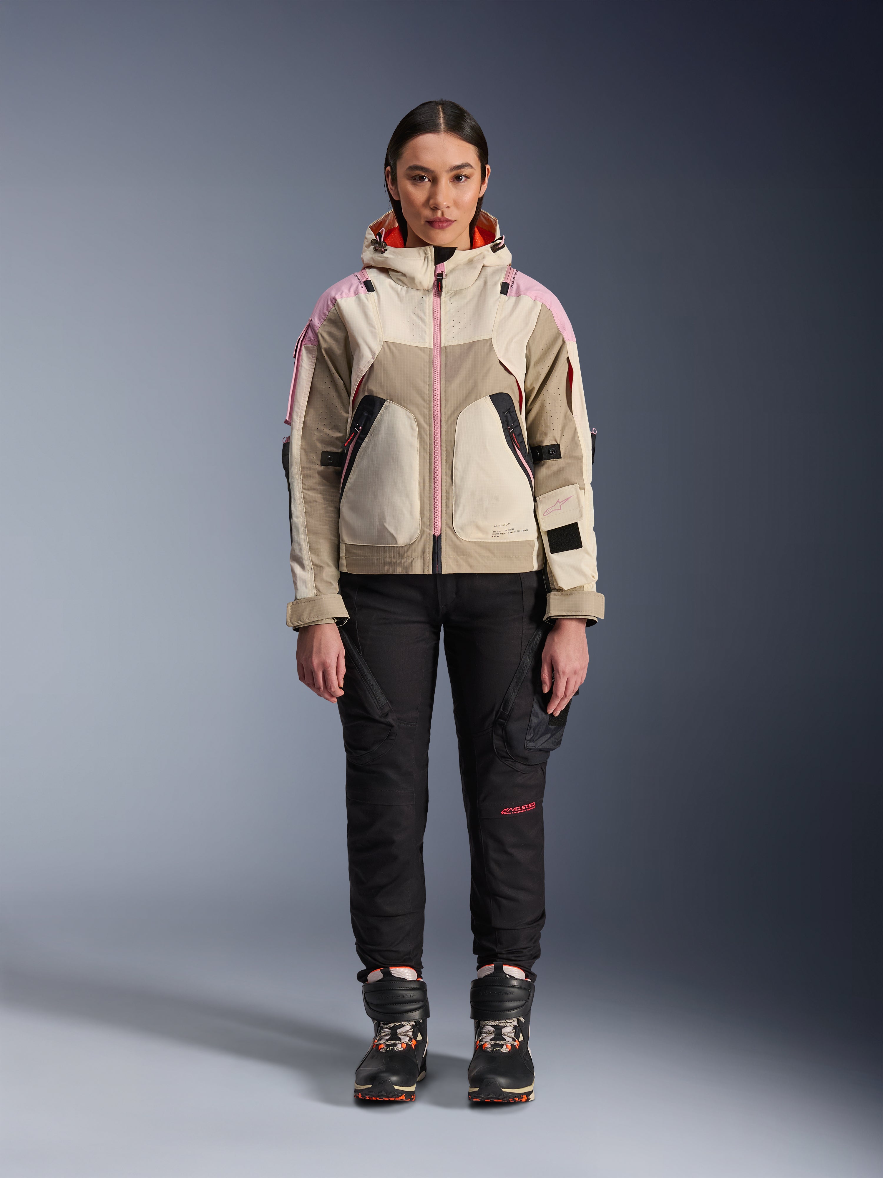 Female model wearing the Alpinestars Molly Air Jacket in Sand Pink Almond, paired with black technical riding pants and boots, front full-body view, urban motorcycle jacket with utility-inspired design and pink accents