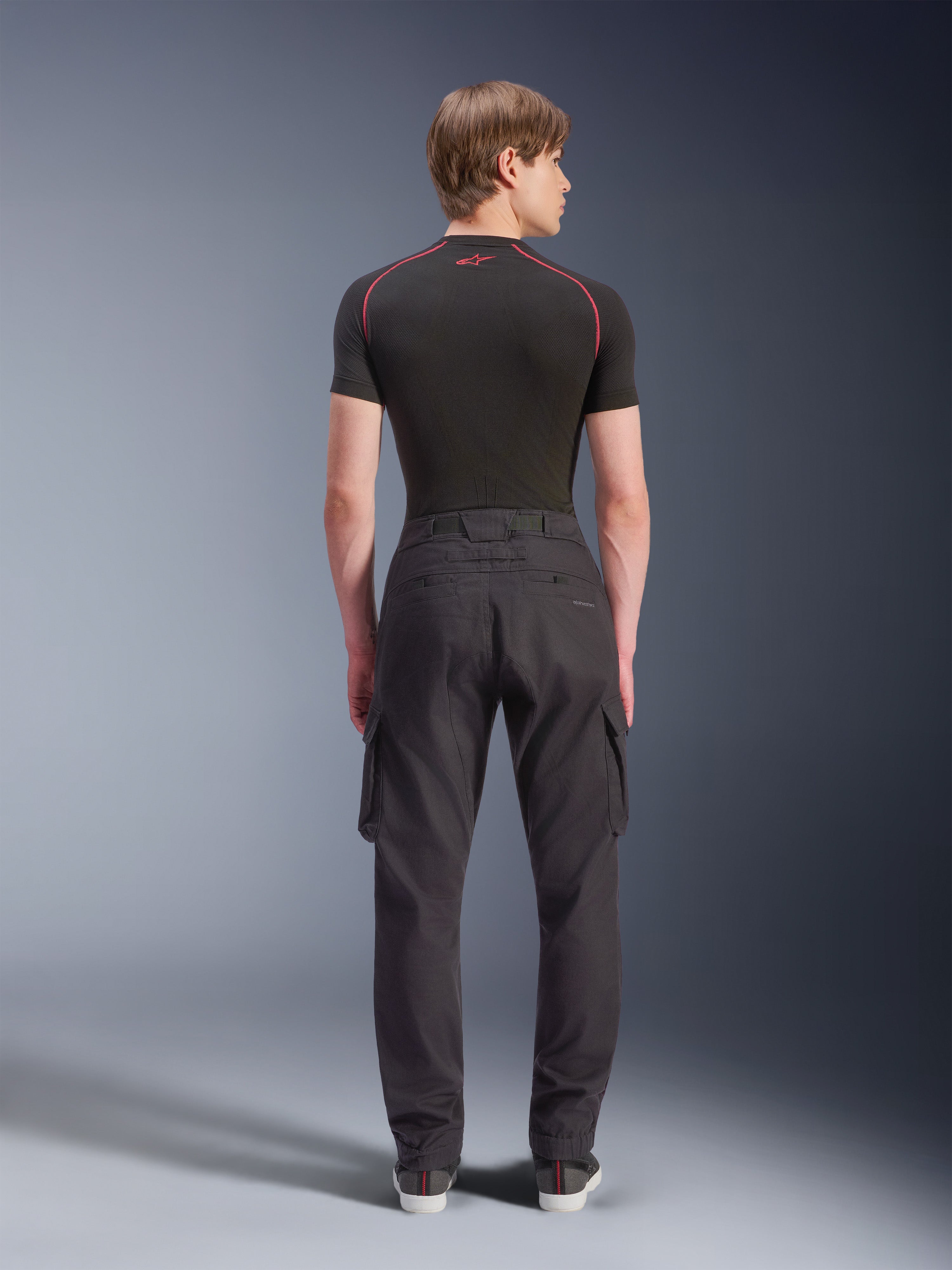 Calças Cargo Alpinestars Flex-AST, calças de motociclismo pretas, vista traseira no modelo, com um corte regular afunilado, bainhas elásticas e bolsos cargo laterais, combinadas com uma base layer preta e sapatilhas casuais