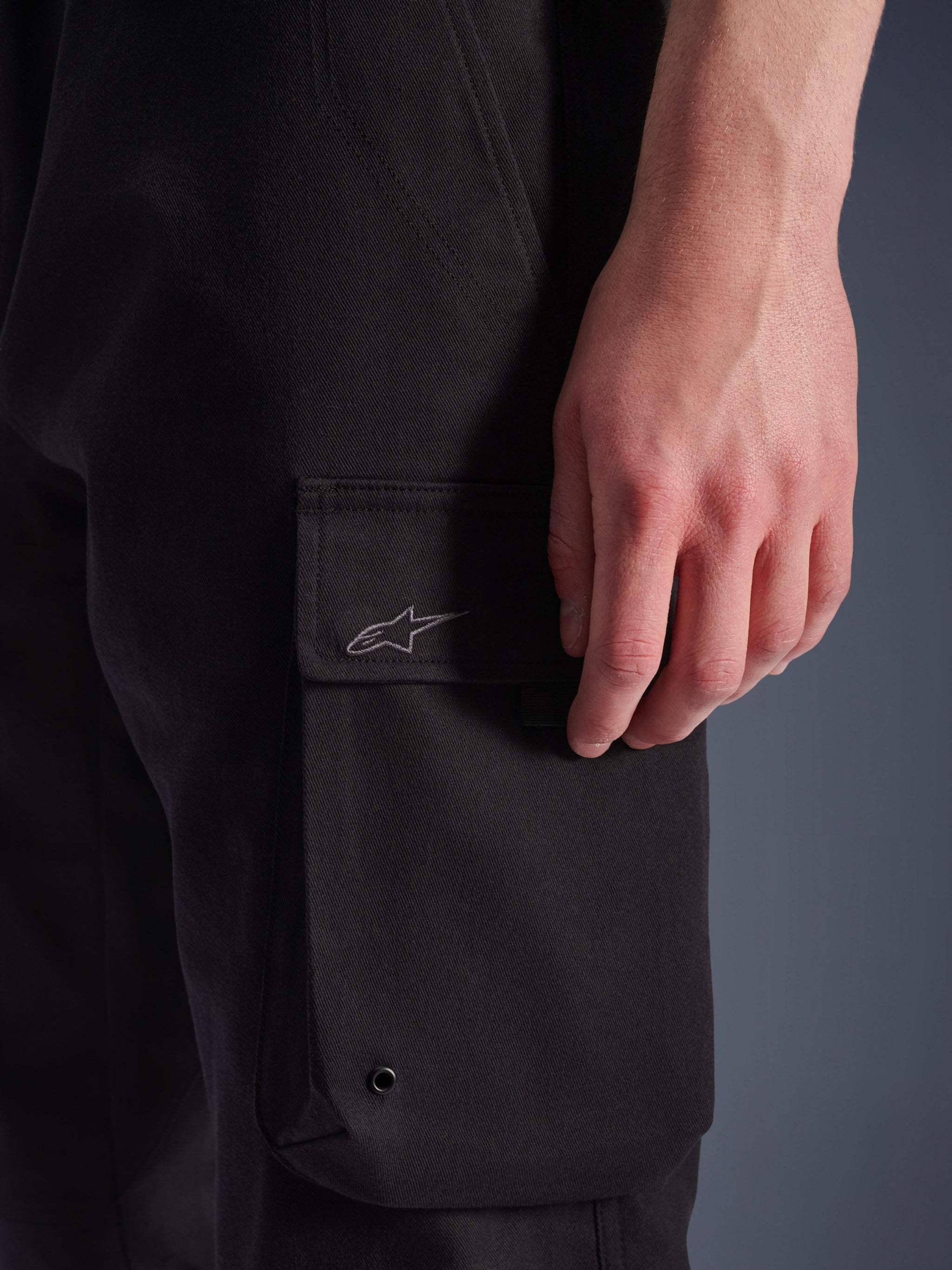 Alpinestars Flex-AST Cargo Pants, Black, close-up detail of side cargo pocket with embroidered Alpinestars logo, durable textile material featuring a metal eyelet, urban motorcycle lifestyle gear