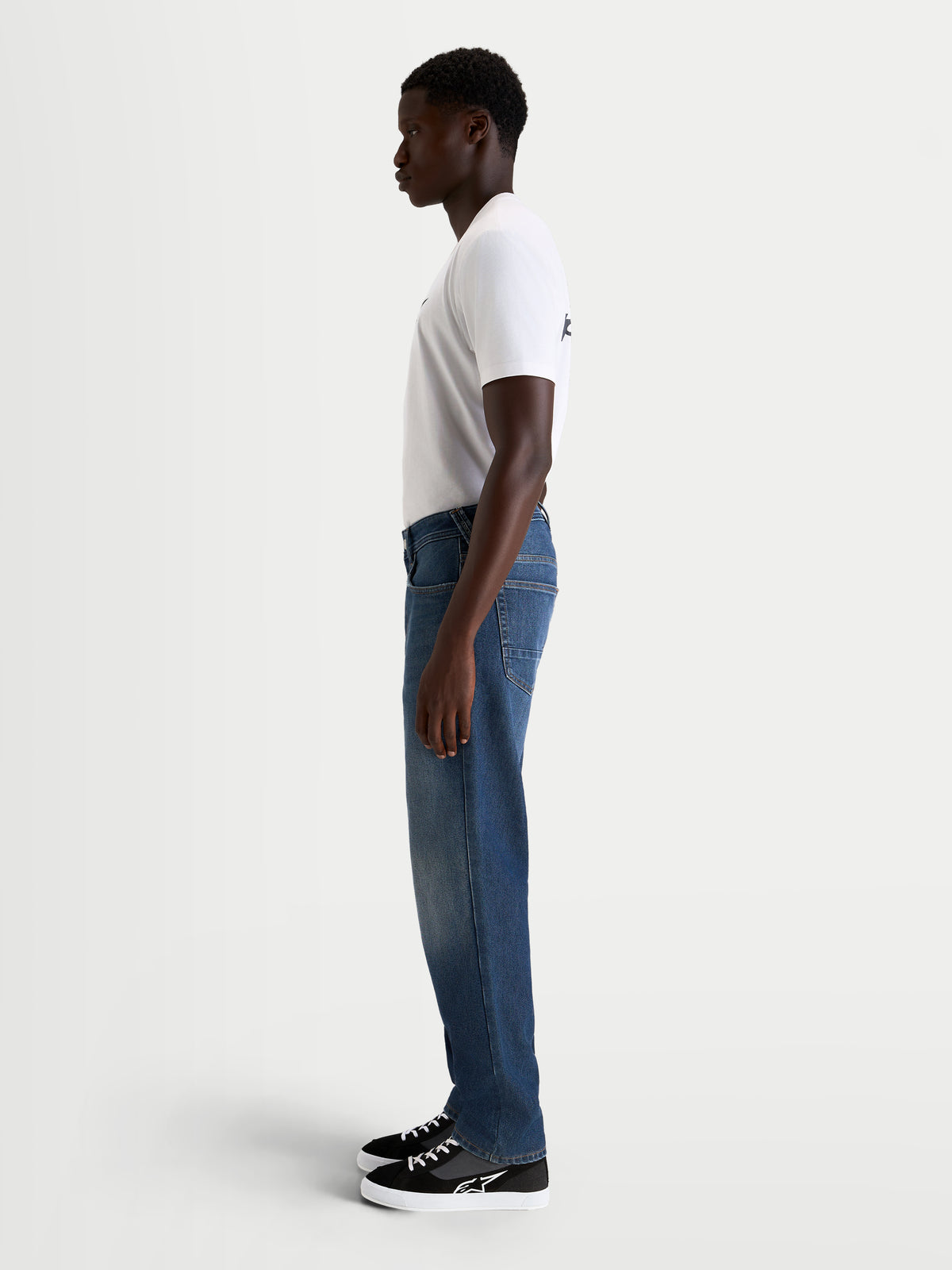 Alpinestars Flex-AST 8 Denim calças de moto urbanas para homem em azul médio, vista de perfil lateral num modelo com t-shirt branca e sapatos de moto casual, ganga de camada única com certificação AA e construção elástica