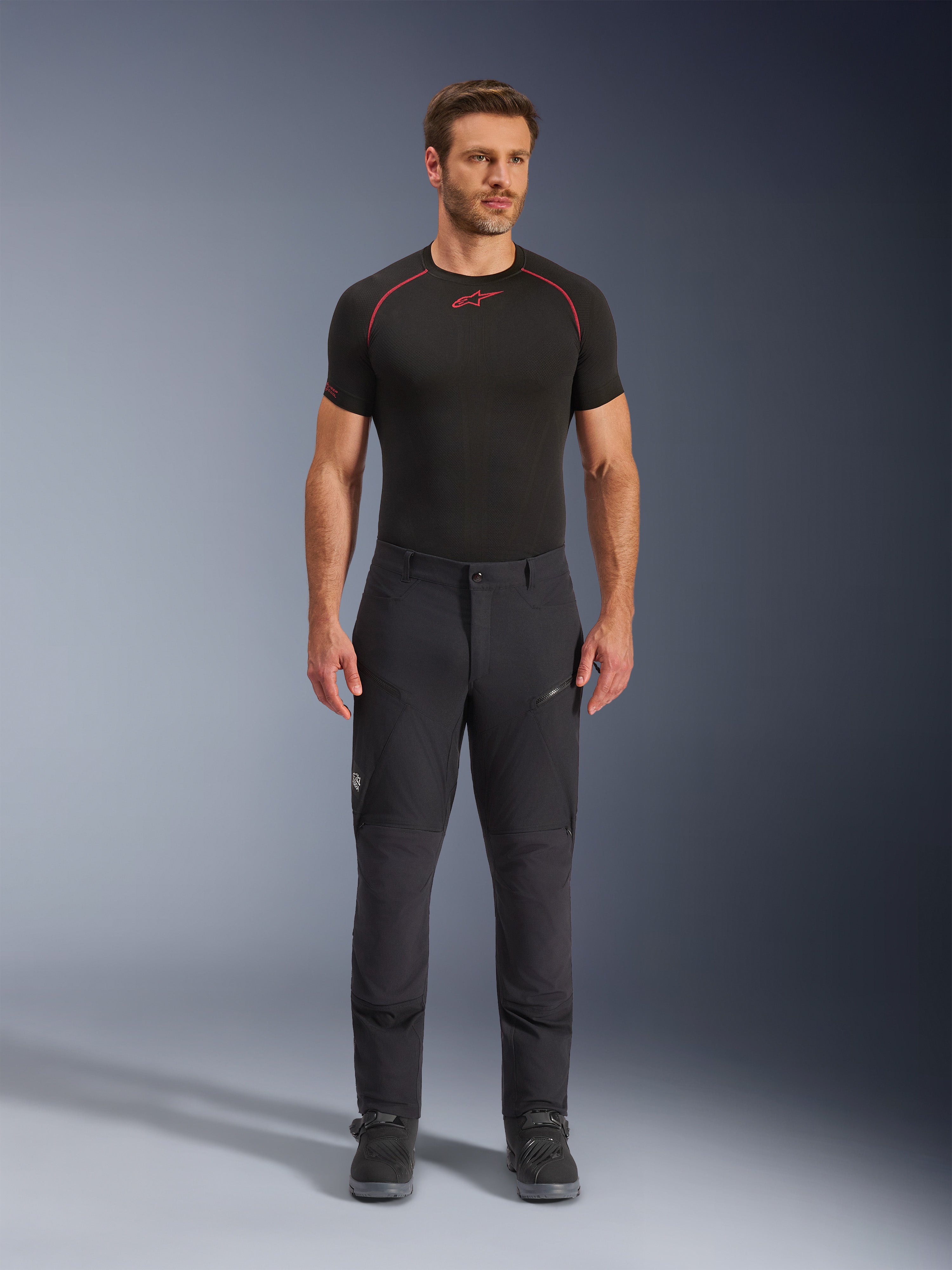 Alpinestars Flex-AST Explorer Pants, calças de motociclista pretas para aventura e touring, homem a usar calças têxteis ajustadas com bolsos cargo com fecho e painéis reforçados, combinadas com uma camada base preta e botas de motociclista, proteção duradoura para condução de aventura