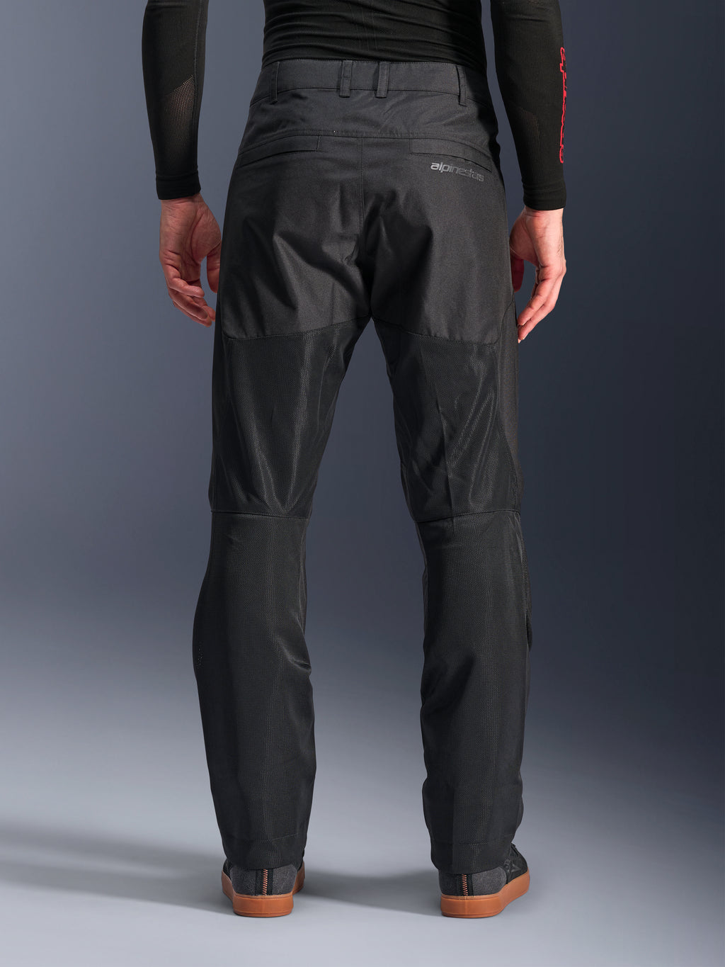 Alpinestars C-1 Air Pants, vista traseira, pretas, calças de motociclismo urbanas em têxtil com grandes painéis de malha técnica na parte de trás das pernas para ventilação, design simples com marca subtil, usadas com uma camada de base preta e sapatilhas de condução casuais.