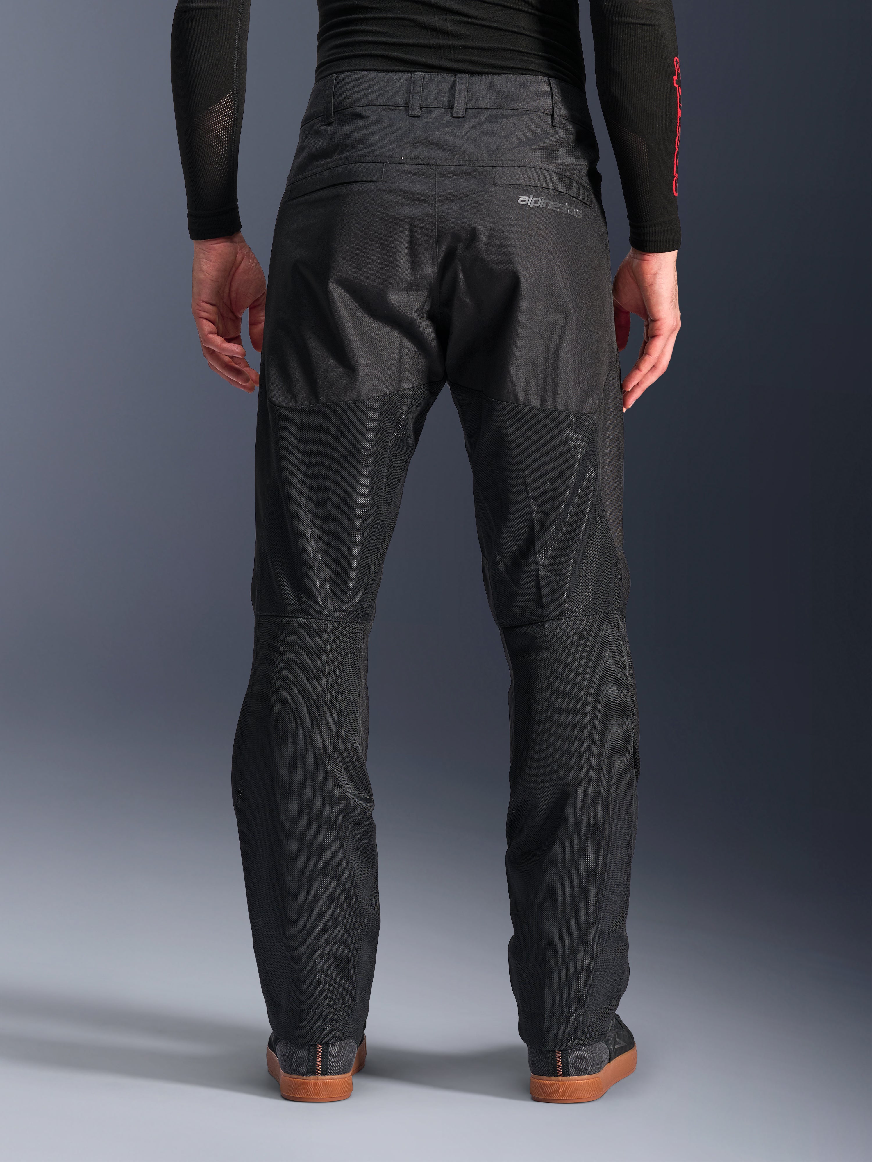Alpinestars C-1 Air Pants, vista traseira, pretas, calças de motociclismo urbanas em têxtil com grandes painéis de malha técnica na parte de trás das pernas para ventilação, design simples com marca subtil, usadas com uma camada de base preta e sapatilhas de condução casuais.