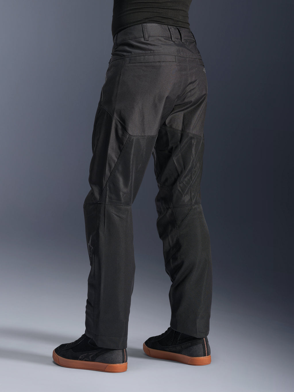 Calças Alpinestars C-1 Air, calças de motociclismo urbanas, pretas, vista traseira mostrando grandes painéis de malha técnica na parte de trás das pernas para ventilação, estilo simples para condução urbana, usadas com ténis pretos casuais.