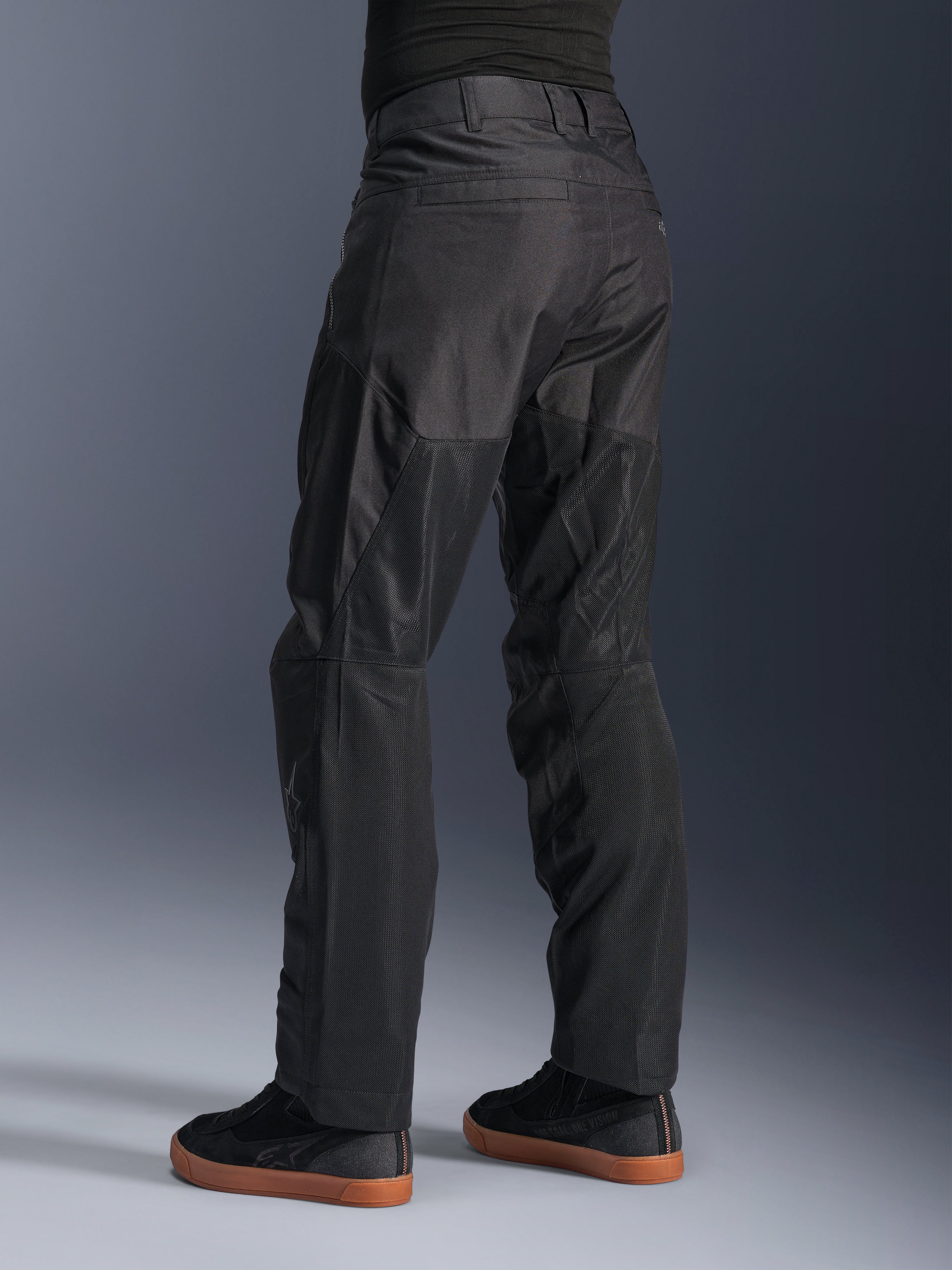 Calças Alpinestars C-1 Air, calças de motociclismo urbanas, pretas, vista traseira mostrando grandes painéis de malha técnica na parte de trás das pernas para ventilação, estilo simples para condução urbana, usadas com ténis pretos casuais.