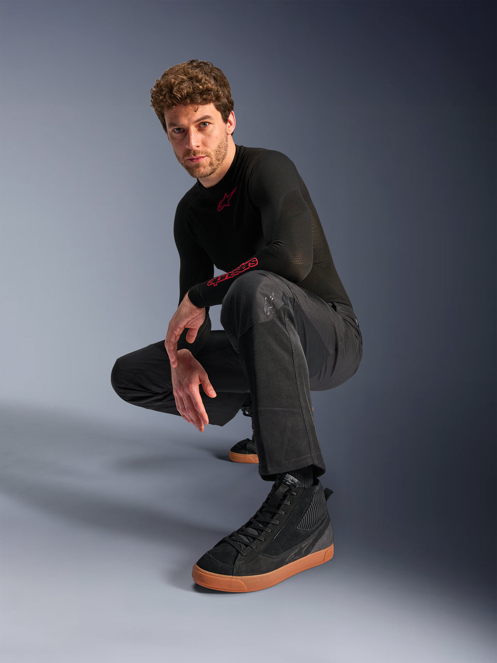 Calças Alpinestars C-1 Air, calças de motociclismo urbanas, pretas, modelo agachado num cenário de estúdio, usadas com uma camisola preta de manga comprida e ténis pretos com sola bege, com painéis de malha respirável e um estilo urbano contemporâneo