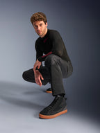 Calças Alpinestars C-1 Air, calças de motociclismo urbanas, pretas, modelo agachado num cenário de estúdio, usadas com uma camisola preta de manga comprida e ténis pretos com sola bege, com painéis de malha respirável e um estilo urbano contemporâneo
