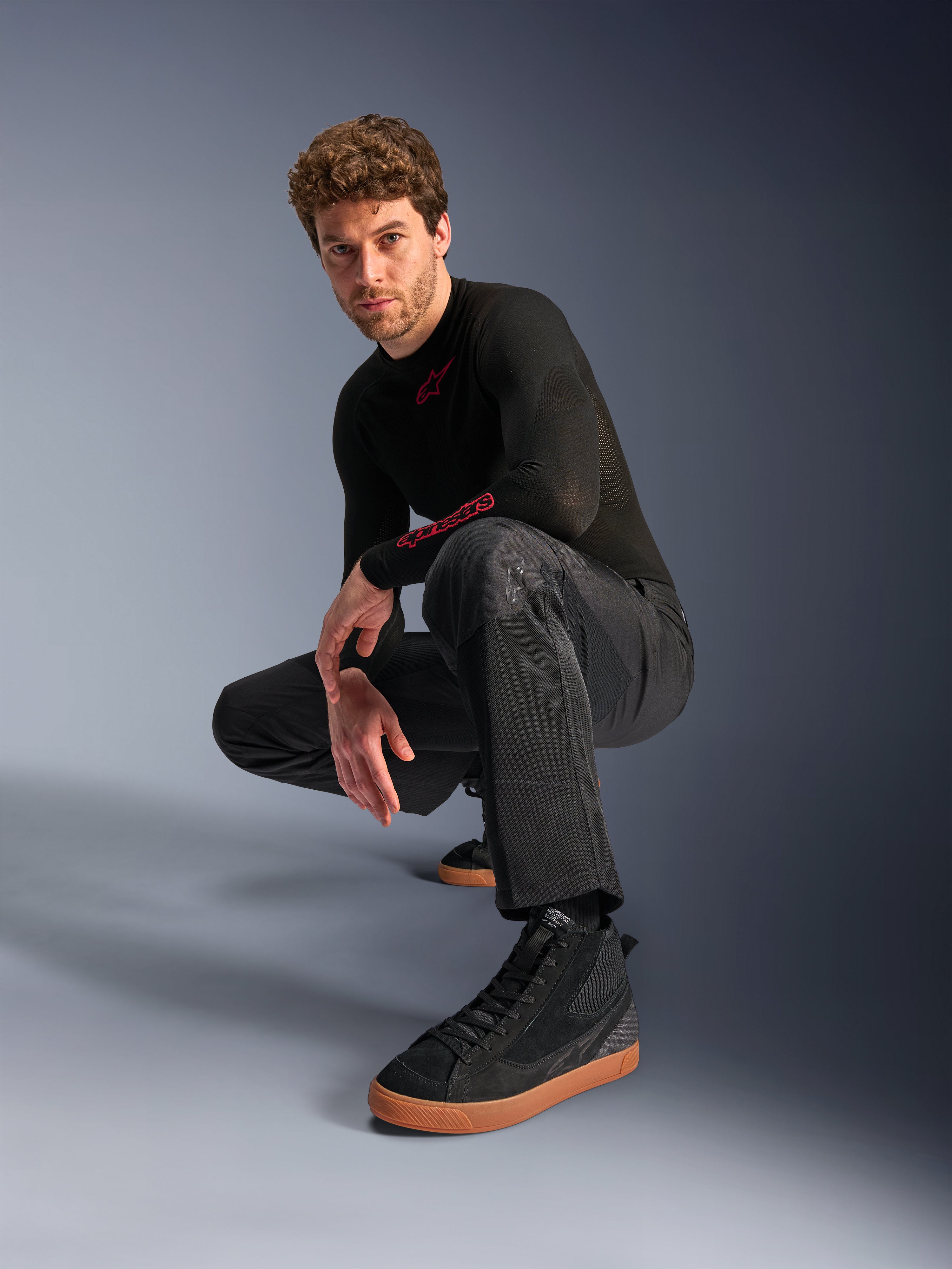 Calças Alpinestars C-1 Air, calças de motociclismo urbanas, pretas, modelo agachado num cenário de estúdio, usadas com uma camisola preta de manga comprida e ténis pretos com sola bege, com painéis de malha respirável e um estilo urbano contemporâneo