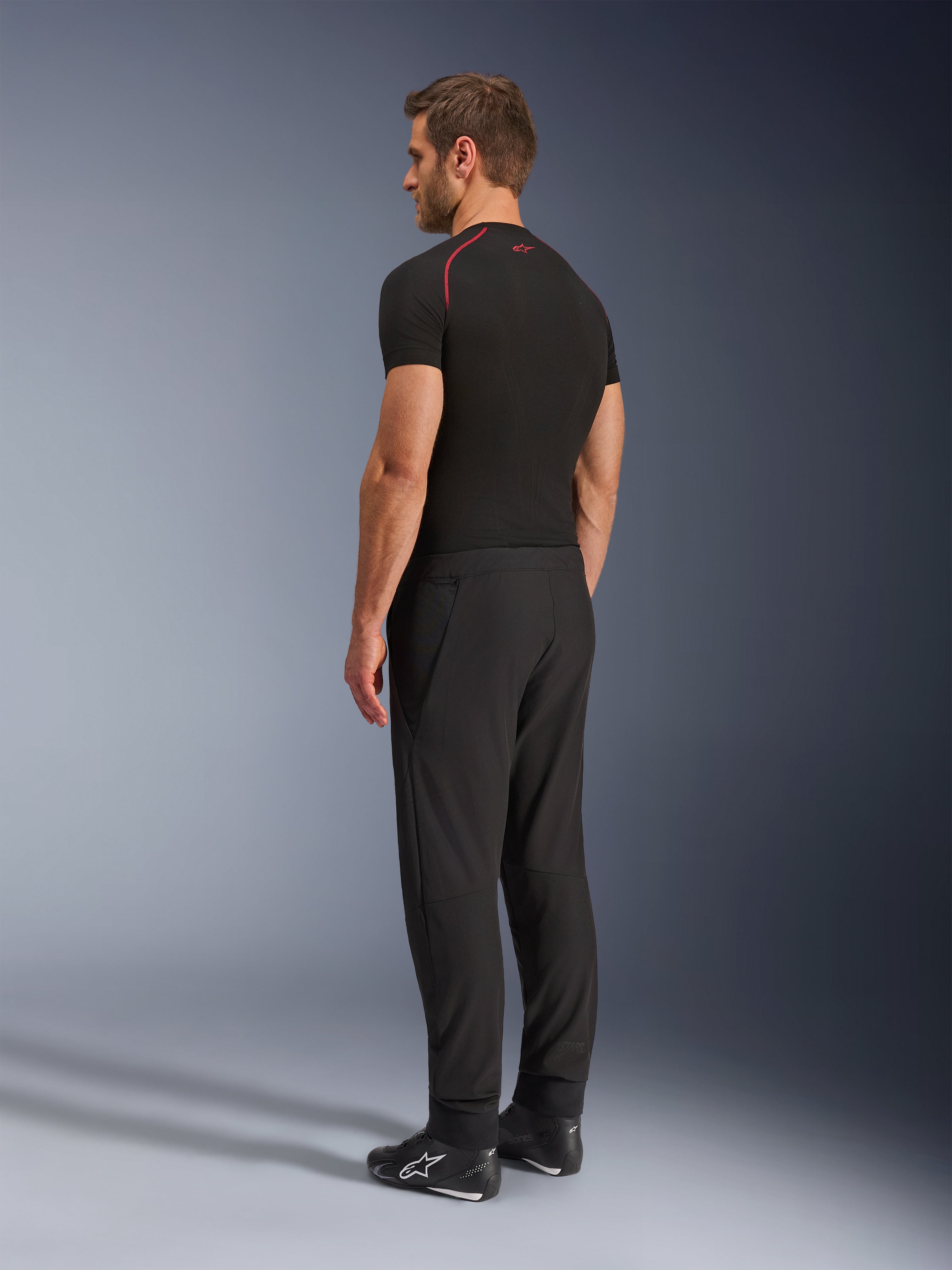 Alpinestars Aeroshell Airflow Pants, calças de motociclismo pretas, tecido softshell leve, usadas com uma t-shirt técnica de manga curta preta e botas de condução pretas, vista de perfil lateral traseira mostrando o corte justo e a construção ergonómica