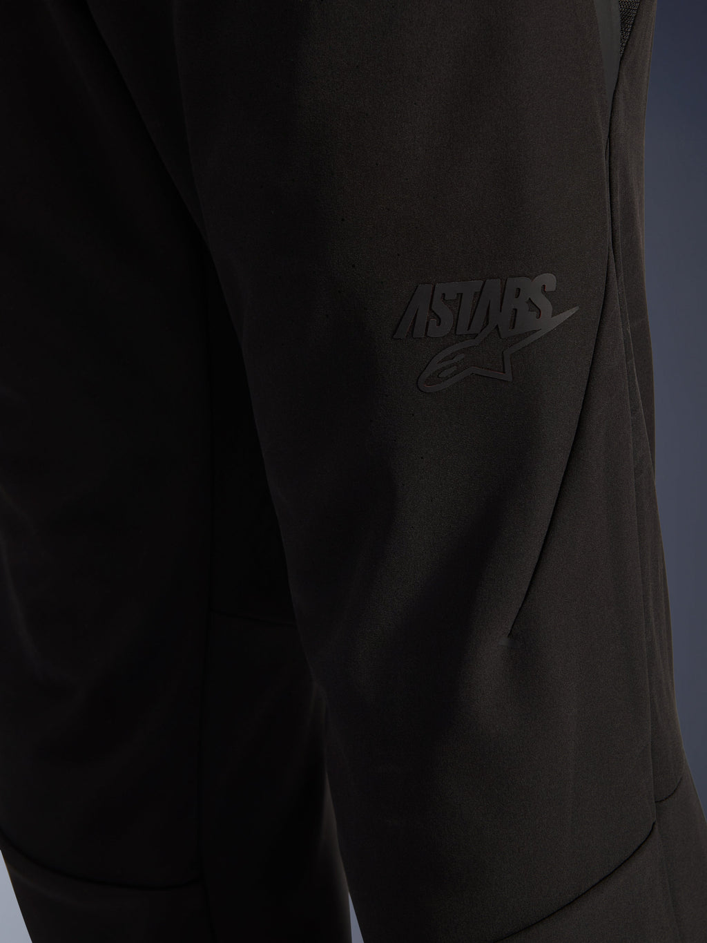 Alpinestars Aeroshell Airflow Pants, calças de motociclismo pretas, vista de pormenor da zona da coxa com logótipo ASTARS preto em relevo, material softshell elástico leve, design de costuras ergonómico