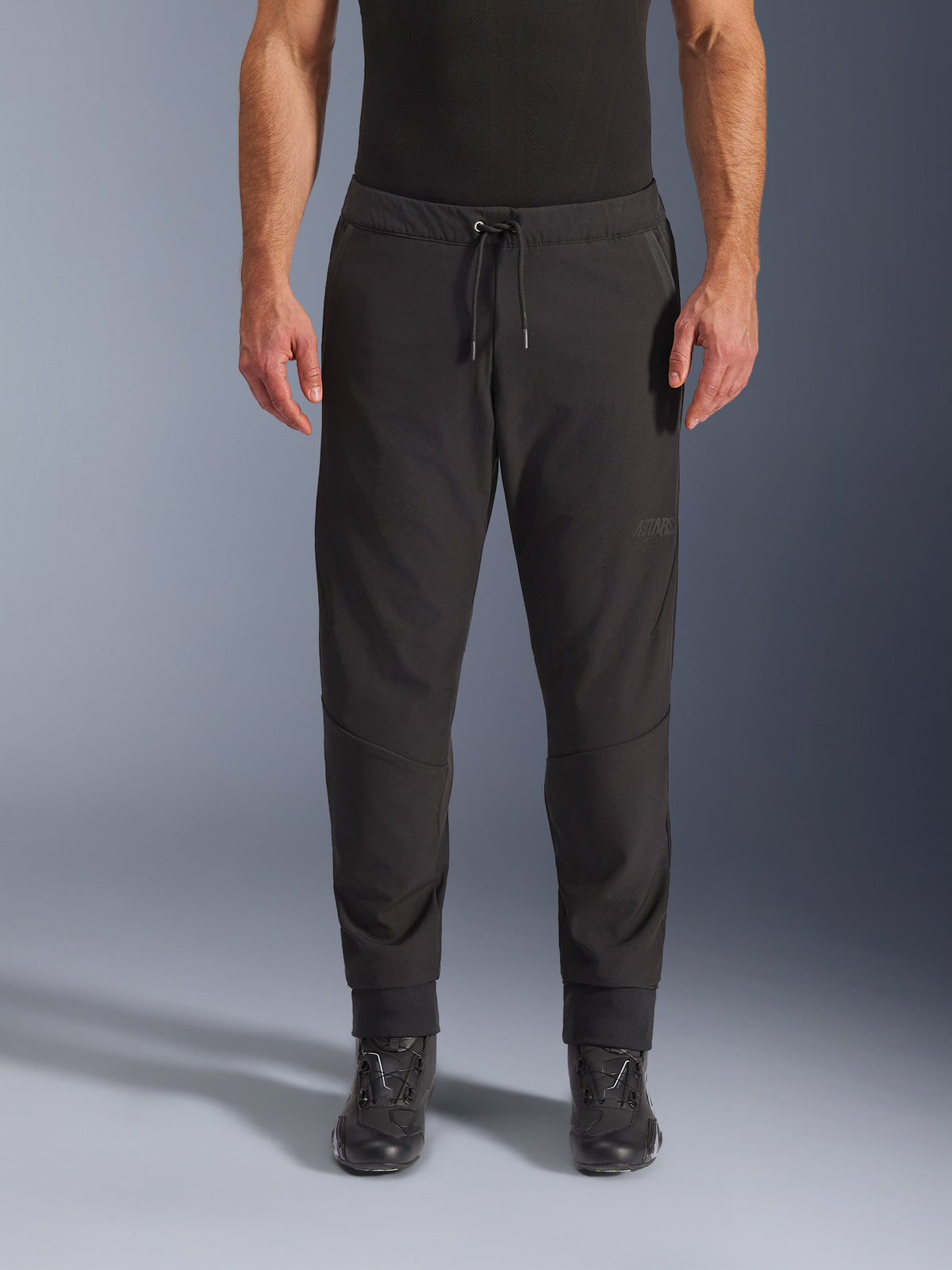 Calças Alpinestars Aeroshell Airflow, calças de motociclismo pretas, vista frontal, usadas com botas de motociclismo pretas, destaque para a cintura com cordão e punhos afunilados nos tornozelos, design softshell leve com logótipo tonal subtil