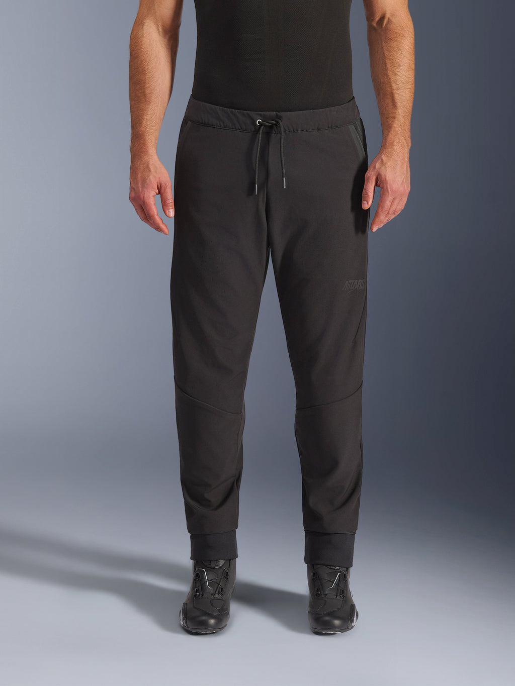 Calças Alpinestars Aeroshell Airflow, calças de motociclismo pretas, vista frontal, usadas com botas de motociclismo pretas, destaque para a cintura com cordão e punhos afunilados nos tornozelos, design softshell leve com logótipo tonal subtil