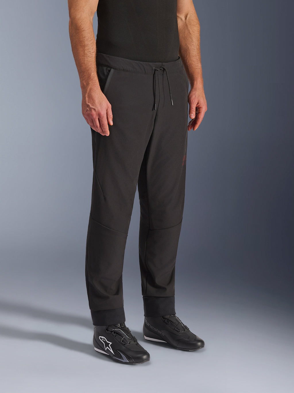 Alpinestars Aeroshell Airflow Pants, calças de motociclismo pretas, usadas por um modelo com calçado de motociclismo preto, com cintura com cordão, punhos nos tornozelos e tecido softshell perfurado a laser para máxima ventilação