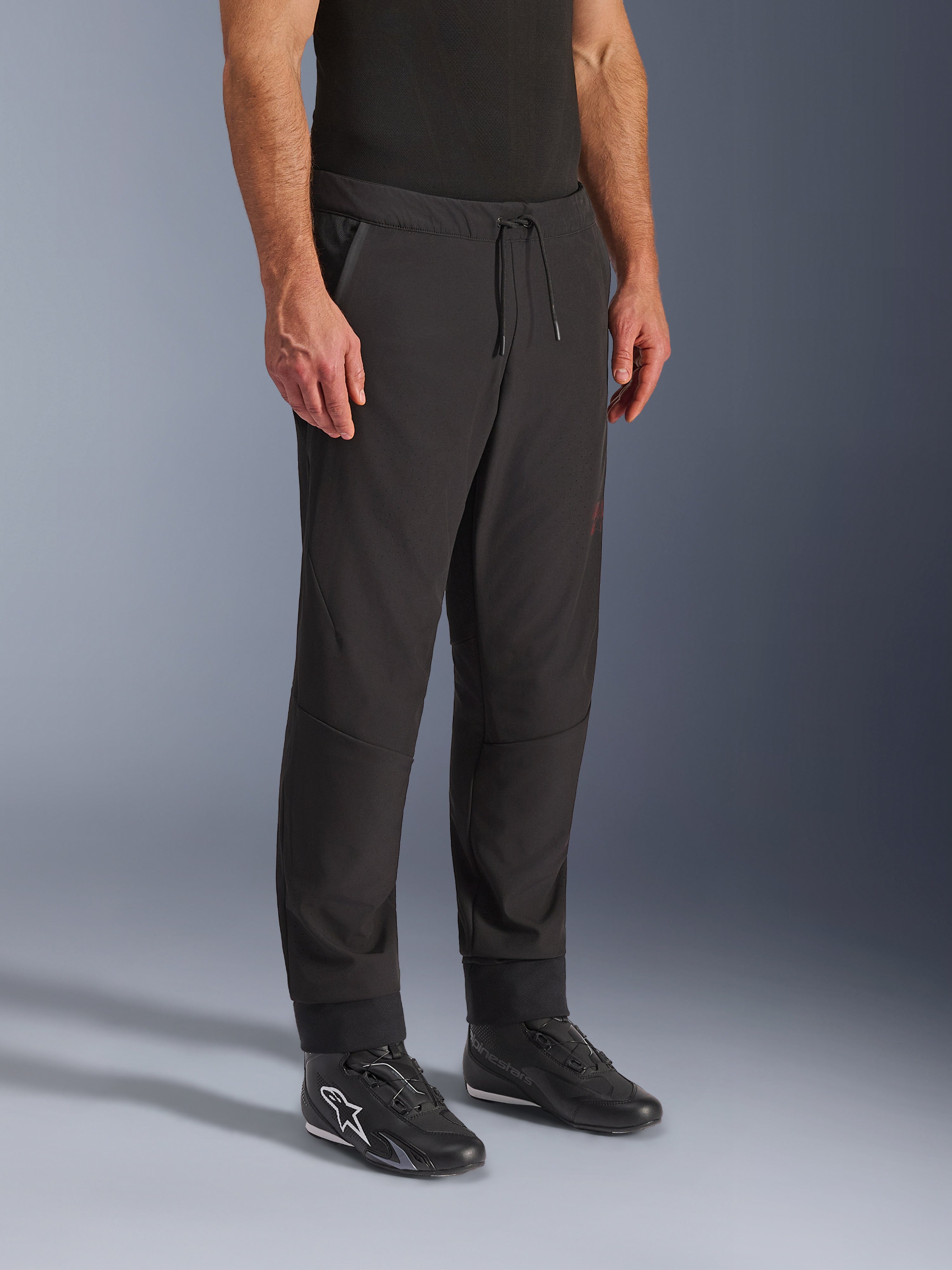 Alpinestars Aeroshell Airflow Pants, calças de motociclismo pretas, usadas por um modelo com calçado de motociclismo preto, com cintura com cordão, punhos nos tornozelos e tecido softshell perfurado a laser para máxima ventilação
