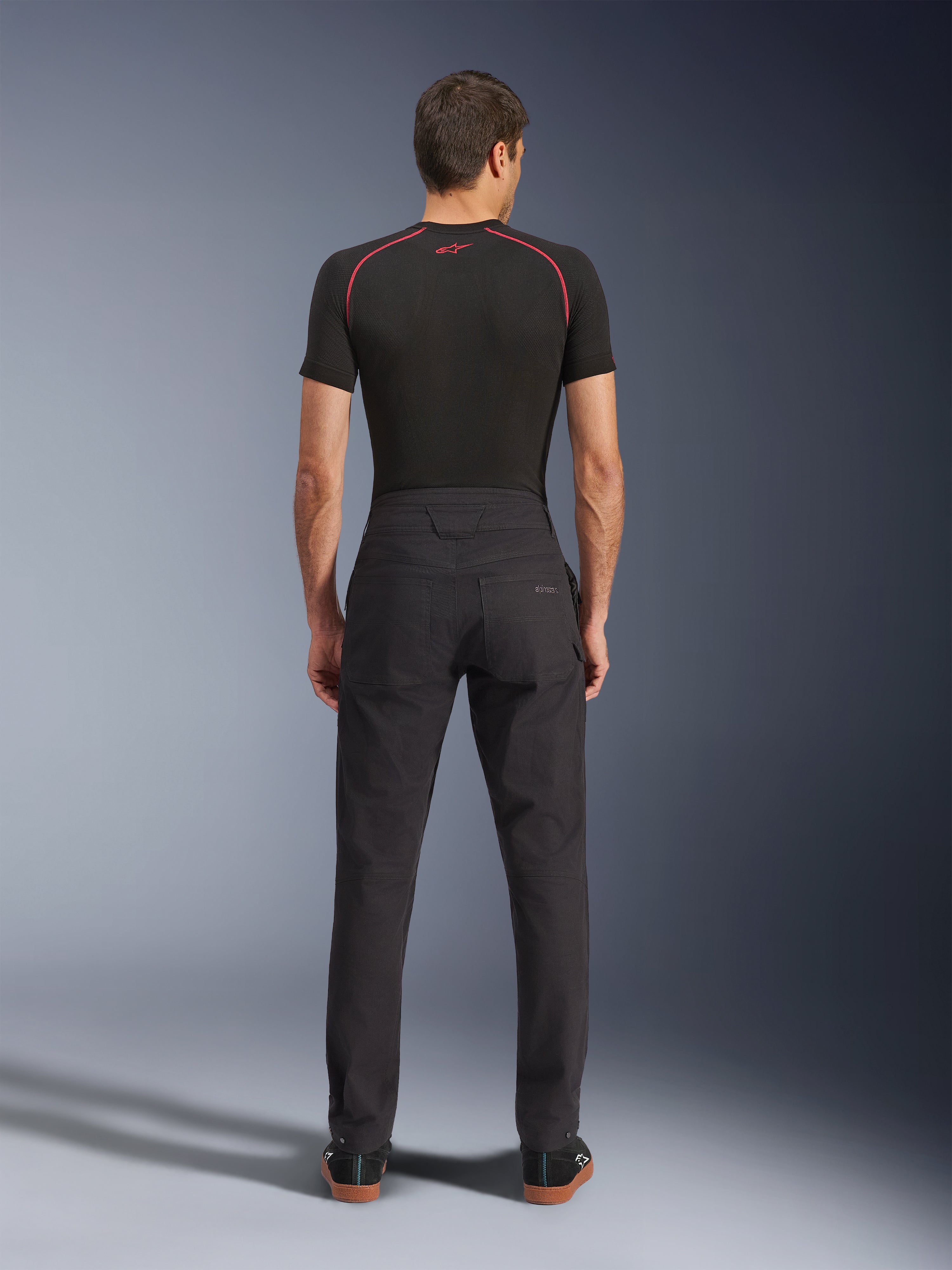 Alpinestars Flex-AST Canvas Pants, calças de motociclismo urbanas, pretas, vista traseira que mostra o design inspirado em roupa de trabalho com bolsos traseiros e subida traseira prolongada, usadas com uma camada de base preta e sapatilhas, corte ergonómico para condução