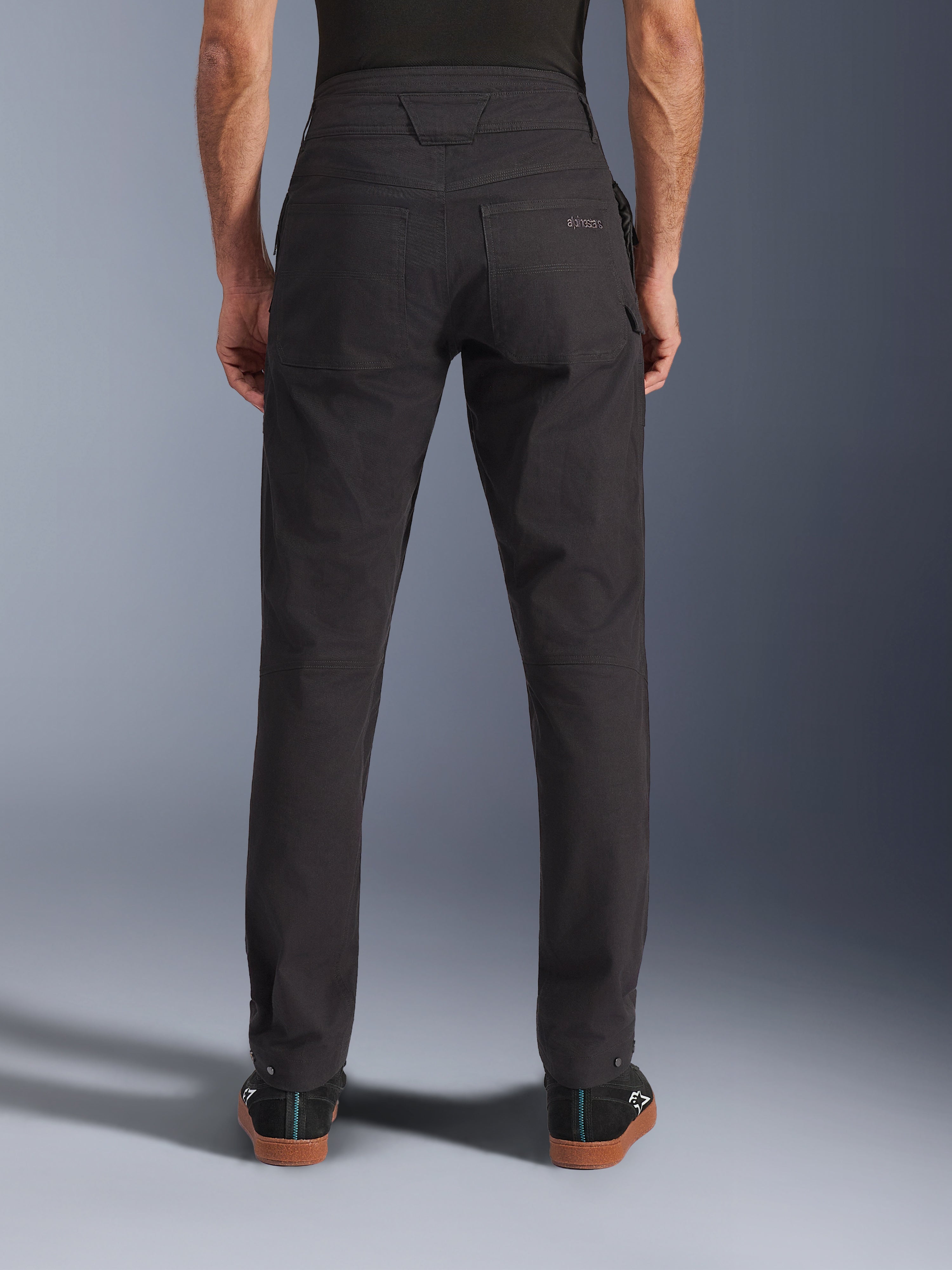 Calças Alpinestars Flex-AST Canvas, calças de motociclismo urbanas, pretas, vista traseira mostrando os bolsos traseiros com logótipo bordado, subida traseira prolongada e costuras ergonómicas nas pernas, usadas com calçado preto casual
