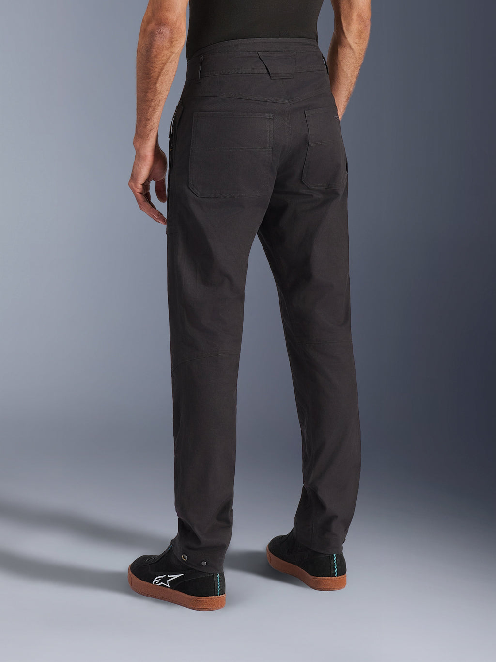 Calças Alpinestars Flex-AST Canvas, pretas, vista traseira mostrando o design clássico de roupa de trabalho com bolsos traseiros grandes, corte ergonómico para condução urbana, usadas com sapatilhas Alpinestars pretas.