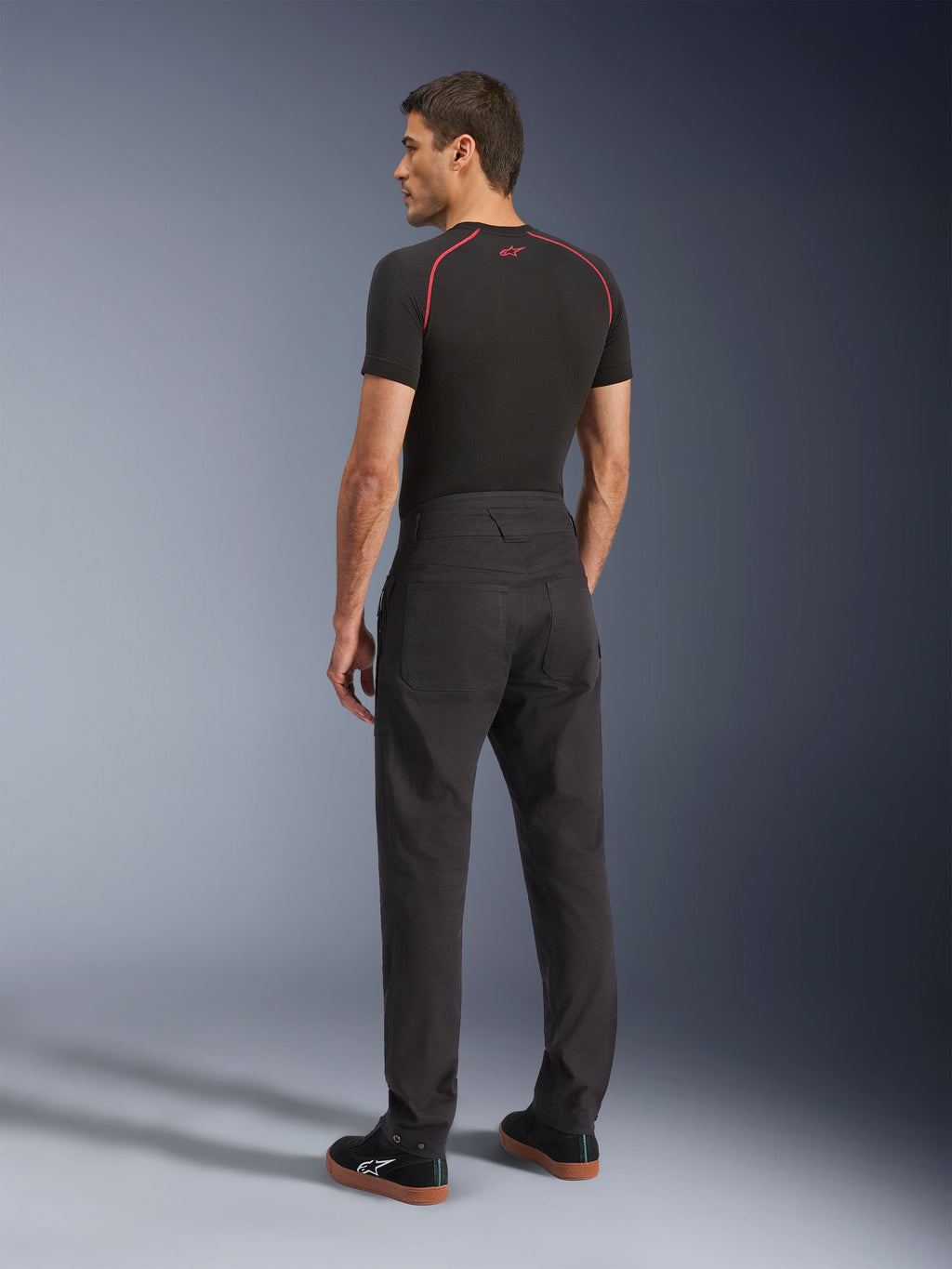 Calças Alpinestars Flex-AST Canvas, calças urbanas de motociclismo pretas, vista traseira num modelo, combinadas com uma camisola de manga curta preta e sapatilhas pretas, com design inspirado em roupa de trabalho, grandes bolsos traseiros e um corte de perna reta moderno.