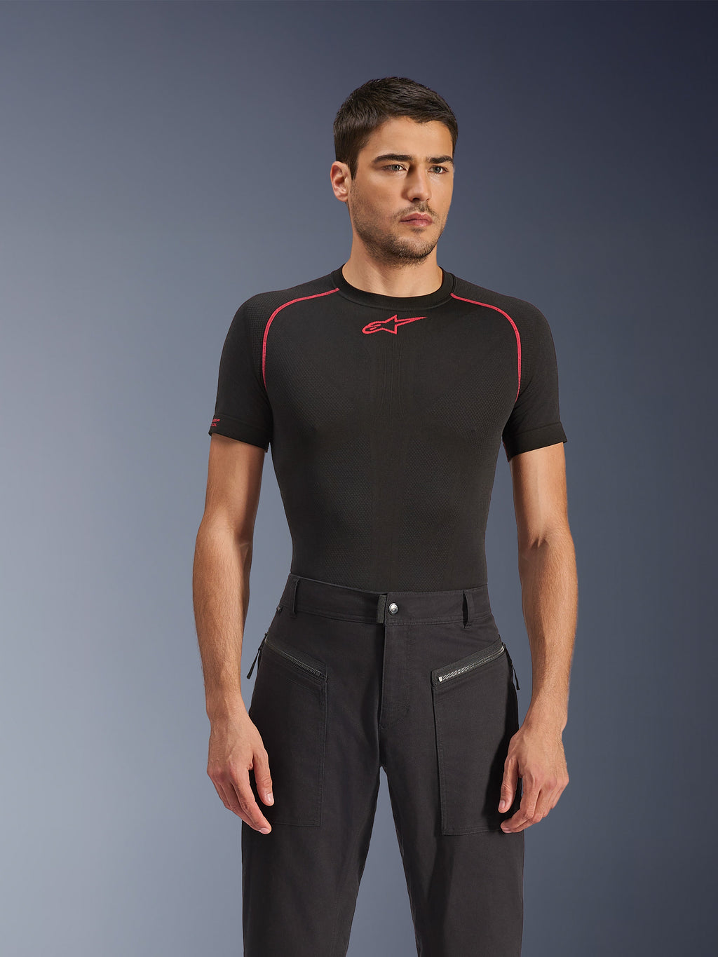 Modelo masculino a usar calças Alpinestars Flex-AST Canvas em preto, calças de motociclismo urbanas com bolsos frontais com fecho de correr visíveis e um design inspirado em roupa de trabalho, combinadas com uma camisola de base preta com detalhes vermelhos