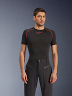 Modelo masculino a usar calças Alpinestars Flex-AST Canvas em preto, calças de motociclismo urbanas com bolsos frontais com fecho de correr visíveis e um design inspirado em roupa de trabalho, combinadas com uma camisola de base preta com detalhes vermelhos