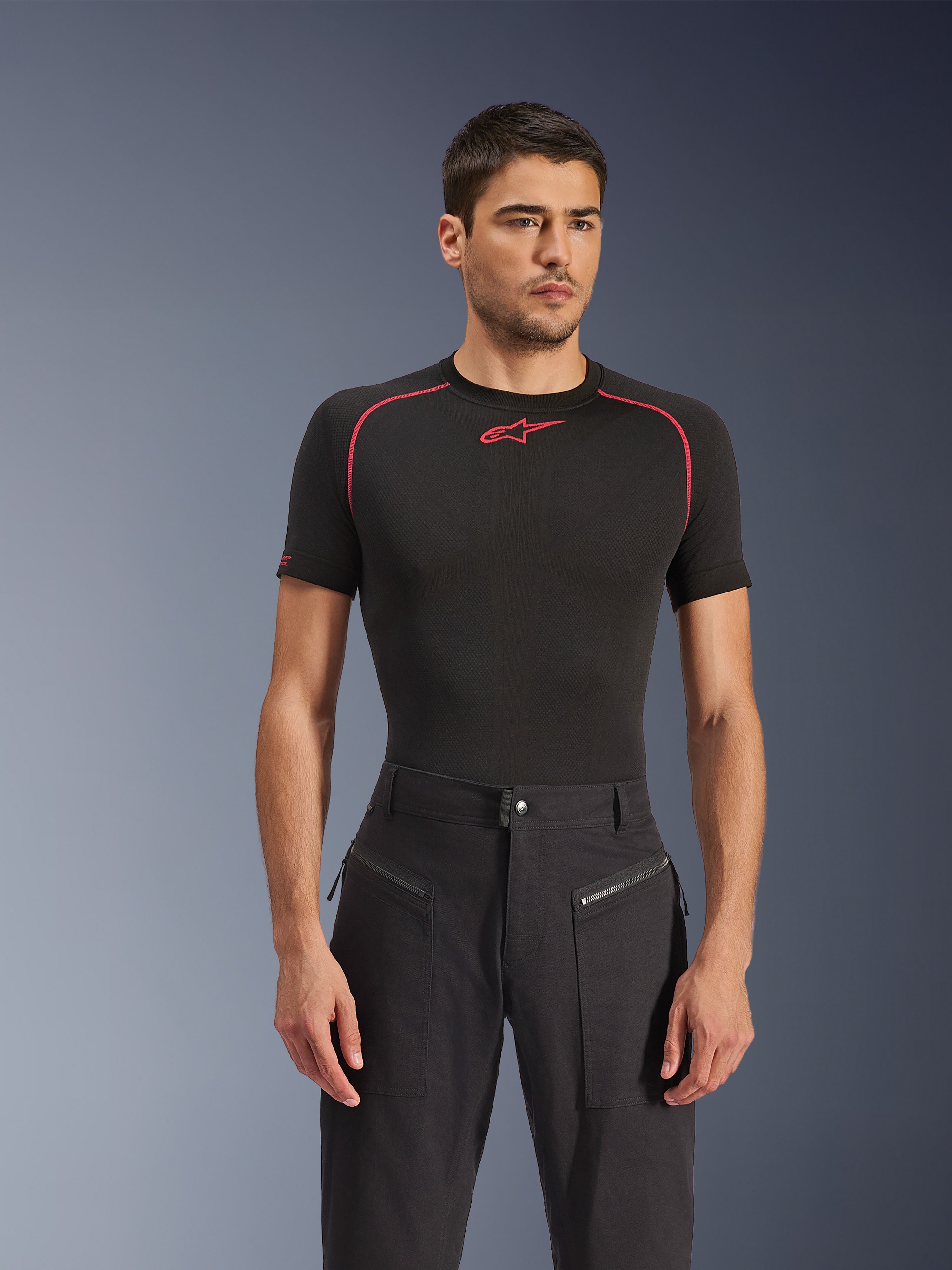 Modelo masculino a usar calças Alpinestars Flex-AST Canvas em preto, calças de motociclismo urbanas com bolsos frontais com fecho de correr visíveis e um design inspirado em roupa de trabalho, combinadas com uma camisola de base preta com detalhes vermelhos
