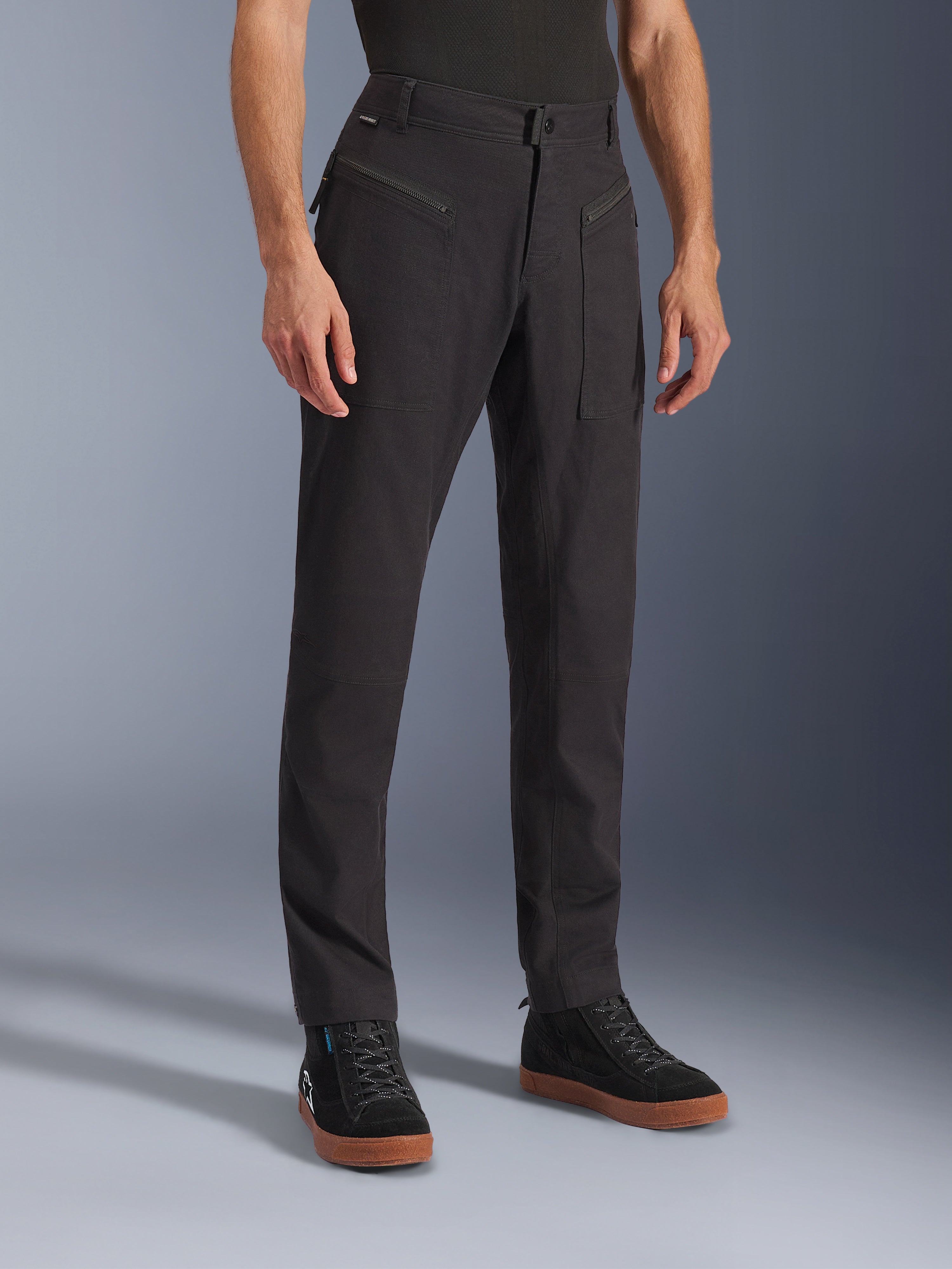 Alpinestars Flex-AST Canvas Pants, calças de motociclismo urbanas, pretas, usadas por um modelo com sapatilhas pretas, com um design clássico de roupa de trabalho, tecido de sarja de policotão elástico e bolsos frontais funcionais com fecho de correr