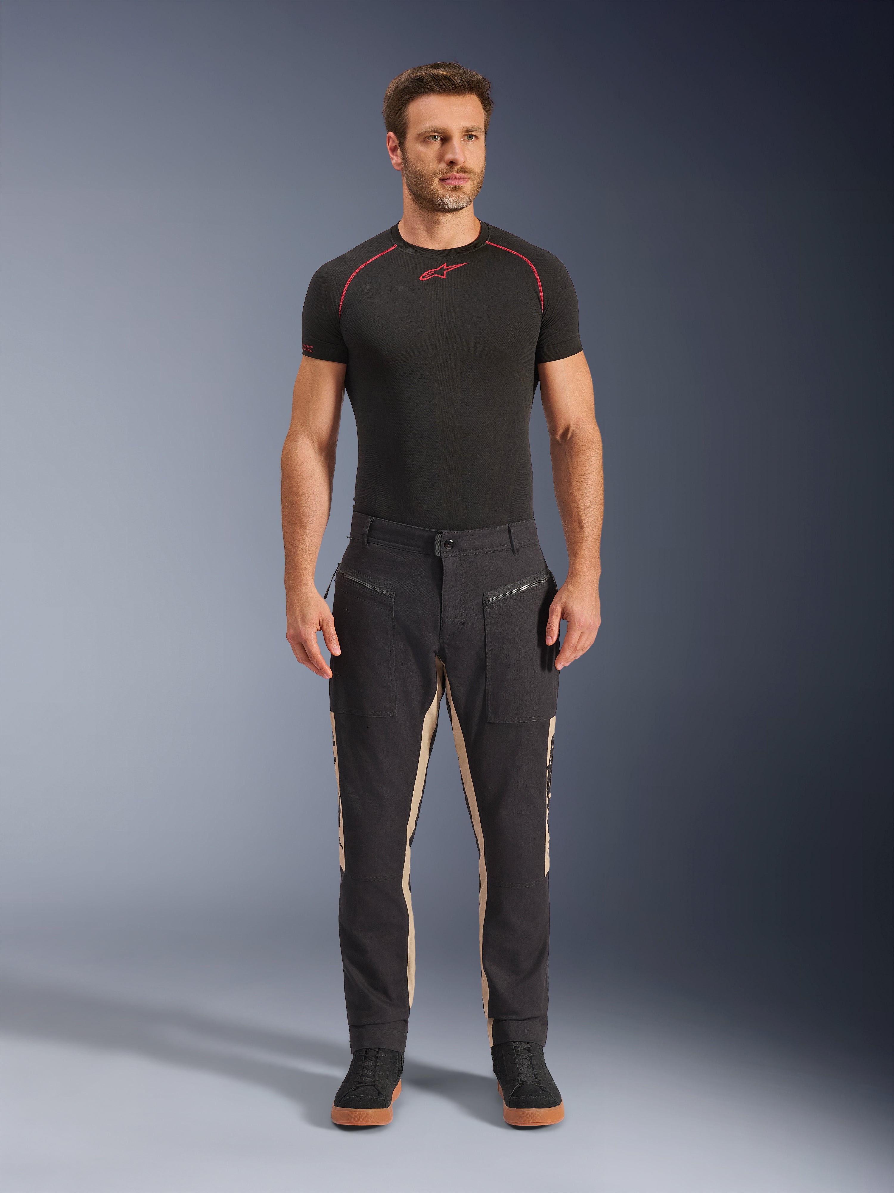 Modelo masculino a usar calças Alpinestars Flex-AST Canvas em Black Aluminum, calças urbanas de motociclismo, pretas com painéis bege, com bolsos utilitários e fechos de correr inspirados em vestuário de trabalho, combinadas com uma camisola interior preta de manga curta e sapatilhas pretas