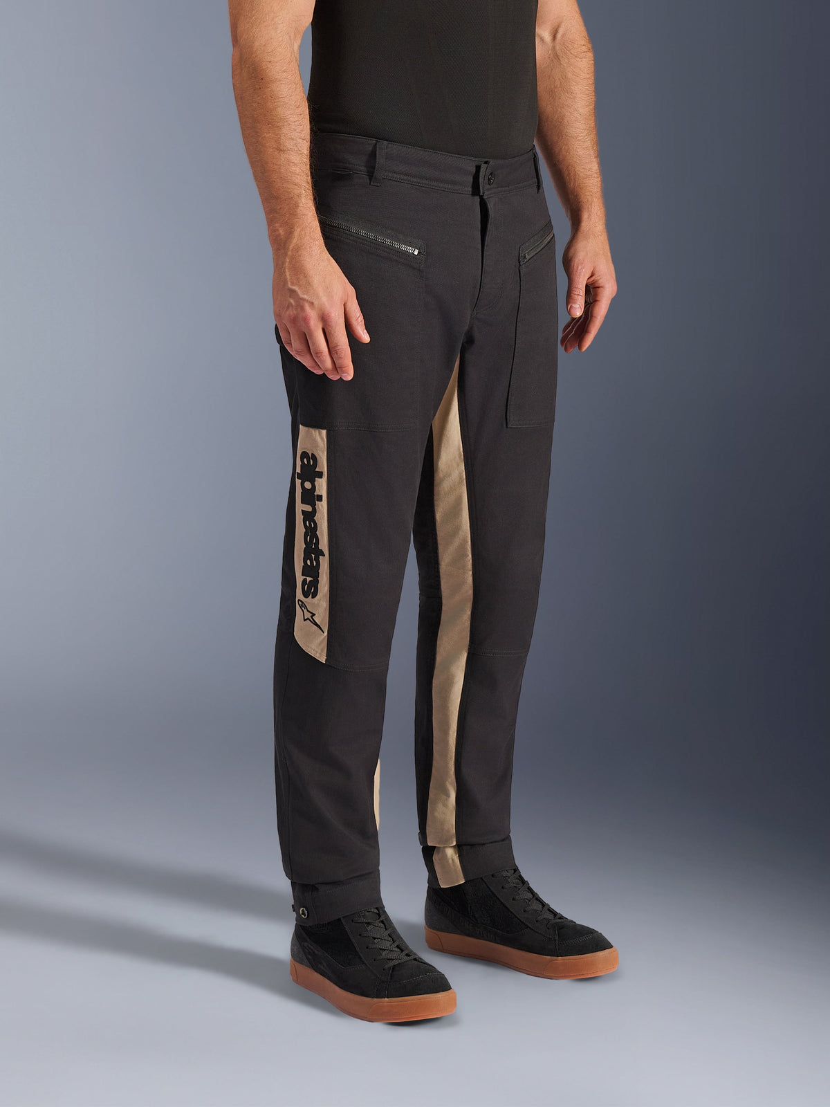 Calças Alpinestars Flex-AST Canvas, calças urbanas de motociclismo, Black Aluminum, com painéis laterais beges com logótipo vertical Alpinestars, design inspirado em vestuário de trabalho com bolsos com fecho e corte ergonómico, usadas com sapatilhas pretas casuais para um visual citadino moderno.