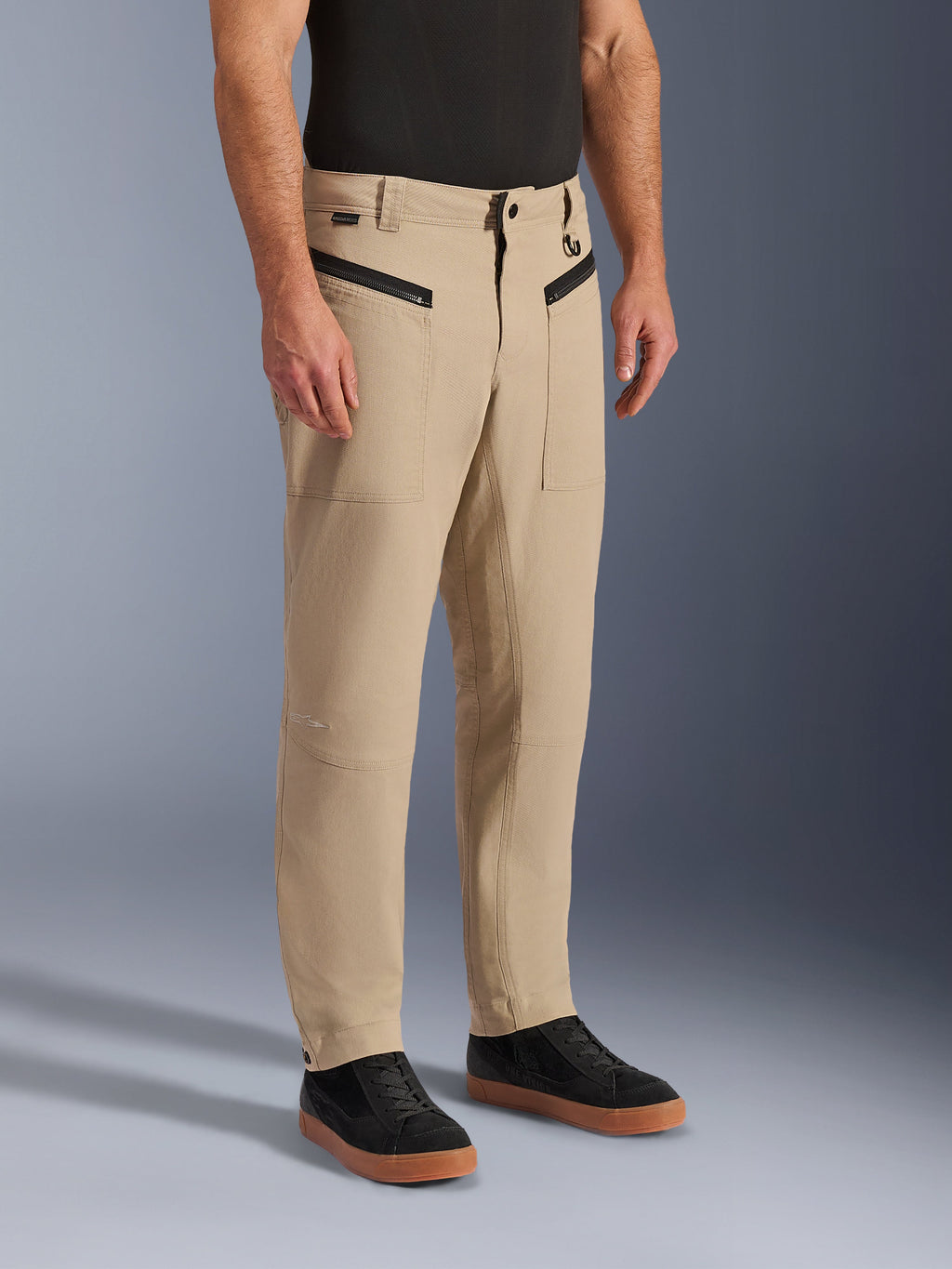 Calças Alpinestars Flex-AST Canvas, calças de motociclismo urbanas, castanho tan, com estilo de roupa de trabalho clássico, bolsos frontais com fecho de correr e painéis ergonómicos nos joelhos, usadas com sapatilhas pretas e uma t-shirt preta, equipamento de condução urbana contemporâneo