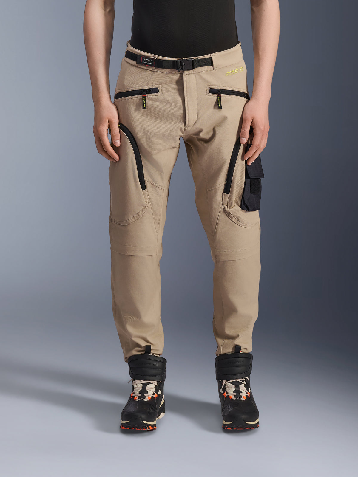 Calças Alpinestars Acteon, calças de motociclismo urbanas, bege areia, com design cargo de vários bolsos, fechos de correr invertidos pretos, cinto integrado e corte afunilado, combinadas com botas pretas e laranja.