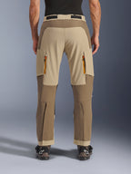 Alpinestars Maxdura Dual Pants, Walnut Aluminum, vista traseira de calças de motociclismo de aventura com painéis bege e em tons de terra, saídas de ventilação com fechos laranja e design por cima da bota, usadas com botas Alpinestars pretas.