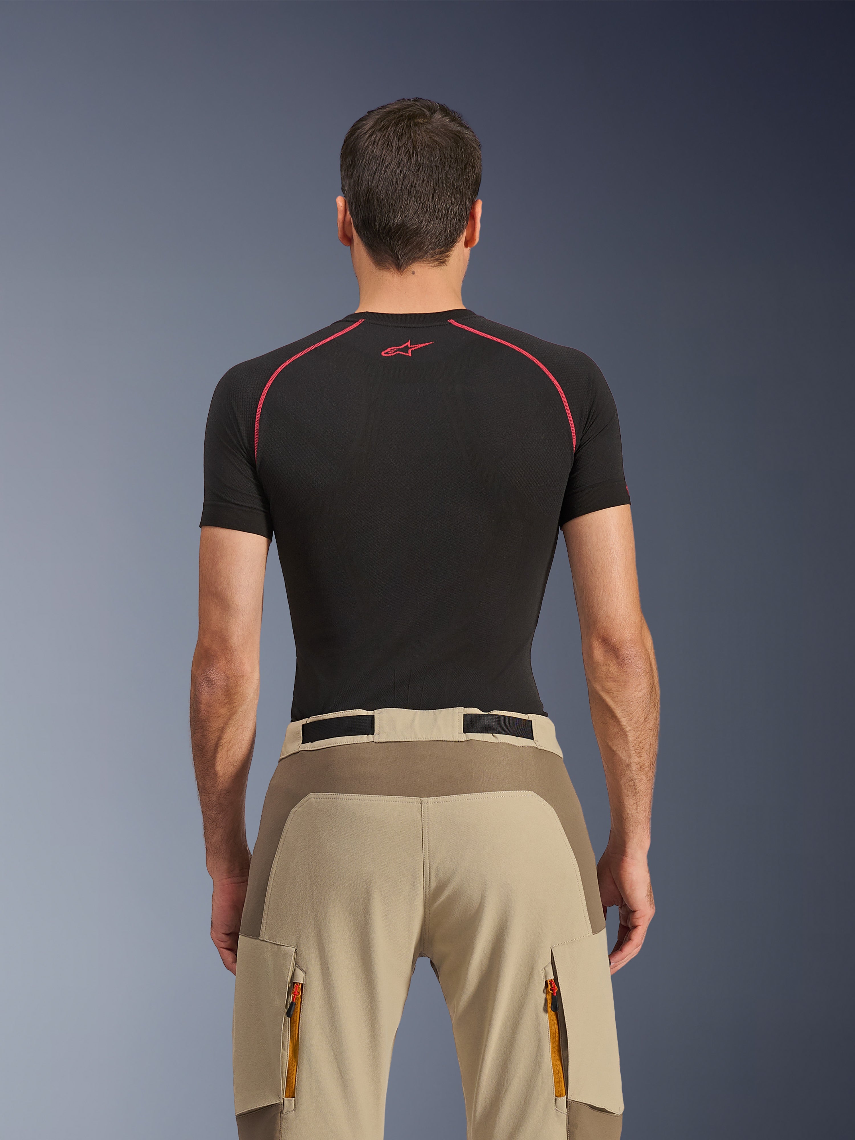 Calças Alpinestars Maxdura Dual, calças de motociclismo de aventura, Walnut Aluminum, vista traseira com painel de assento reforçado e tecido elástico bege, bolsos cargo com fecho de correr e puxadores cor-de-laranja, usadas com uma camisola base layer preta da Alpinestars.