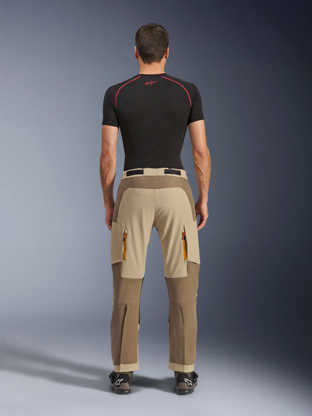 Alpinestars Maxdura Dual Pants em Walnut Aluminum, vista traseira com design em dois tons de castanho e bege, calças de aventura com bolsos cargo com fecho e assento reforçado, usadas com camisola de compressão preta e botas de motociclismo.