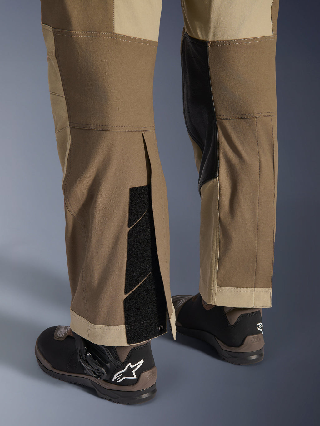 Calças Alpinestars Maxdura Dual em Walnut Aluminum, vista detalhada da parte inferior da perna e da bainha com design por cima da bota, fechos de velcro ajustáveis e painéis interiores reforçados, combinadas com botas de motociclismo pretas.