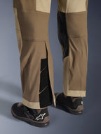 Calças Alpinestars Maxdura Dual em Walnut Aluminum, vista detalhada da parte inferior da perna e da bainha com design por cima da bota, fechos de velcro ajustáveis e painéis interiores reforçados, combinadas com botas de motociclismo pretas.