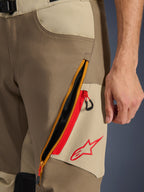 Alpinestars Maxdura Dual Pants, Walnut e Aluminum, pormenor da coxa com um grande sistema de ventilação diagonal com fecho de correr e detalhes vermelhos, logótipo Alpinestars vermelho num painel bege, equipamento de motociclismo de aventura