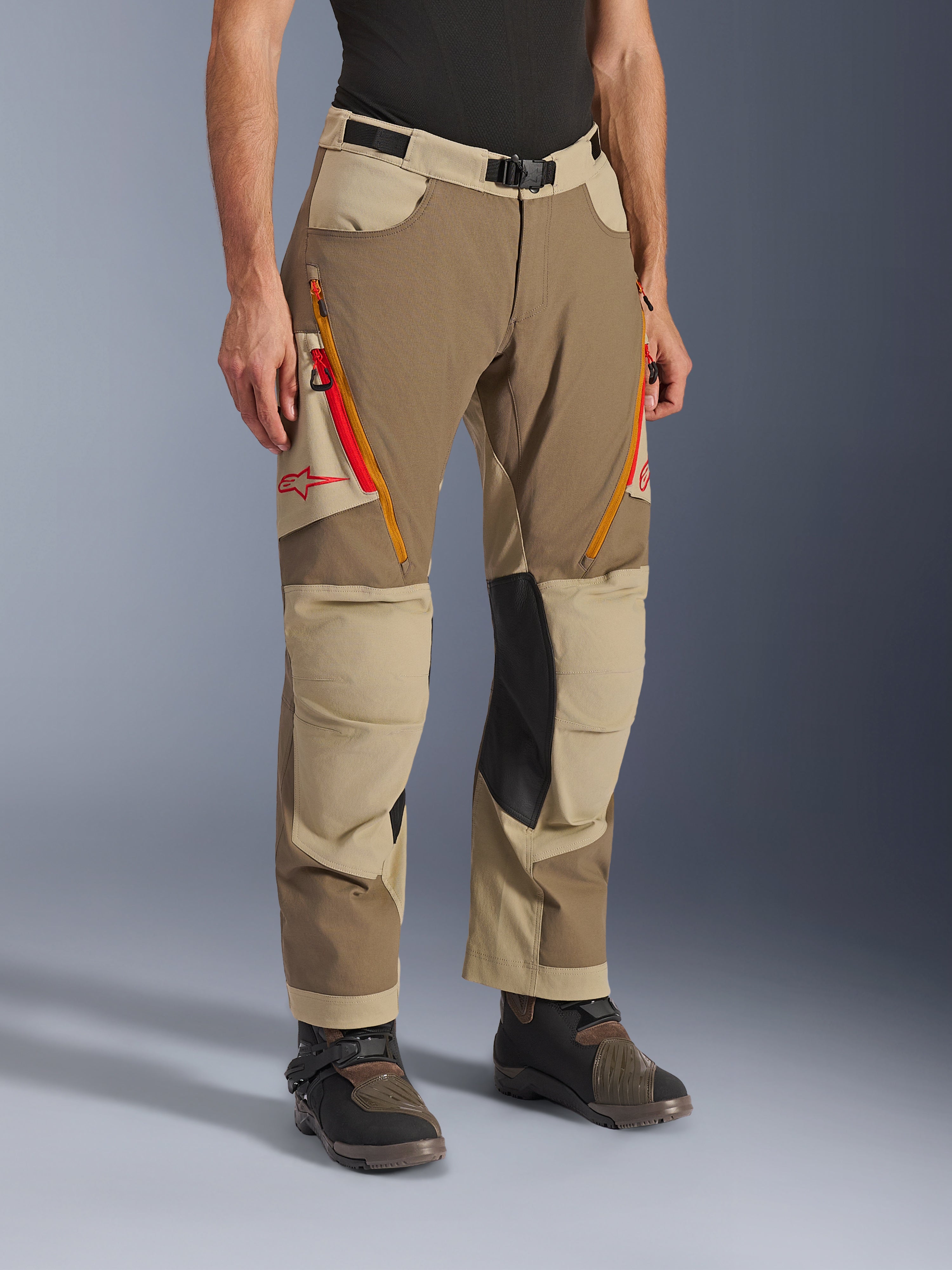 Calças Alpinestars Maxdura Dual, calças de aventura em castanho Walnut Aluminum e caqui, com aberturas de ventilação nas coxas com fecho em laranja e vermelho, painéis interiores dos joelhos em pele preta reforçada e logótipo Astars vermelho, usadas com botas técnicas