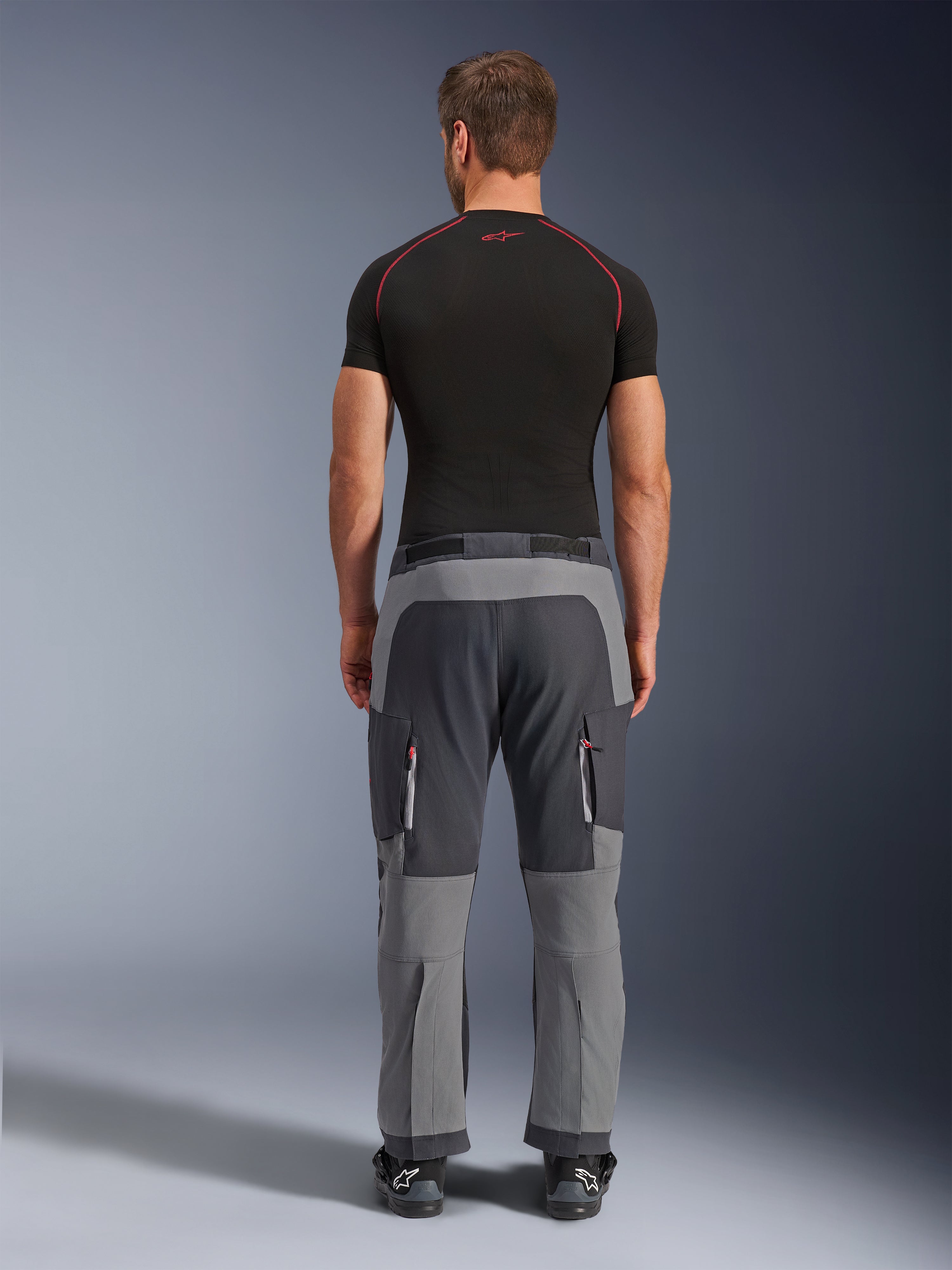 Alpinestars Maxdura Dual Pants, calças de motociclismo de aventura em cinzento escuro e cinzento profundo, vista traseira num modelo a usar t-shirt preta e botas, com painéis cinzentos de vários tons e bolsos cargo com fecho e detalhes vermelhos.