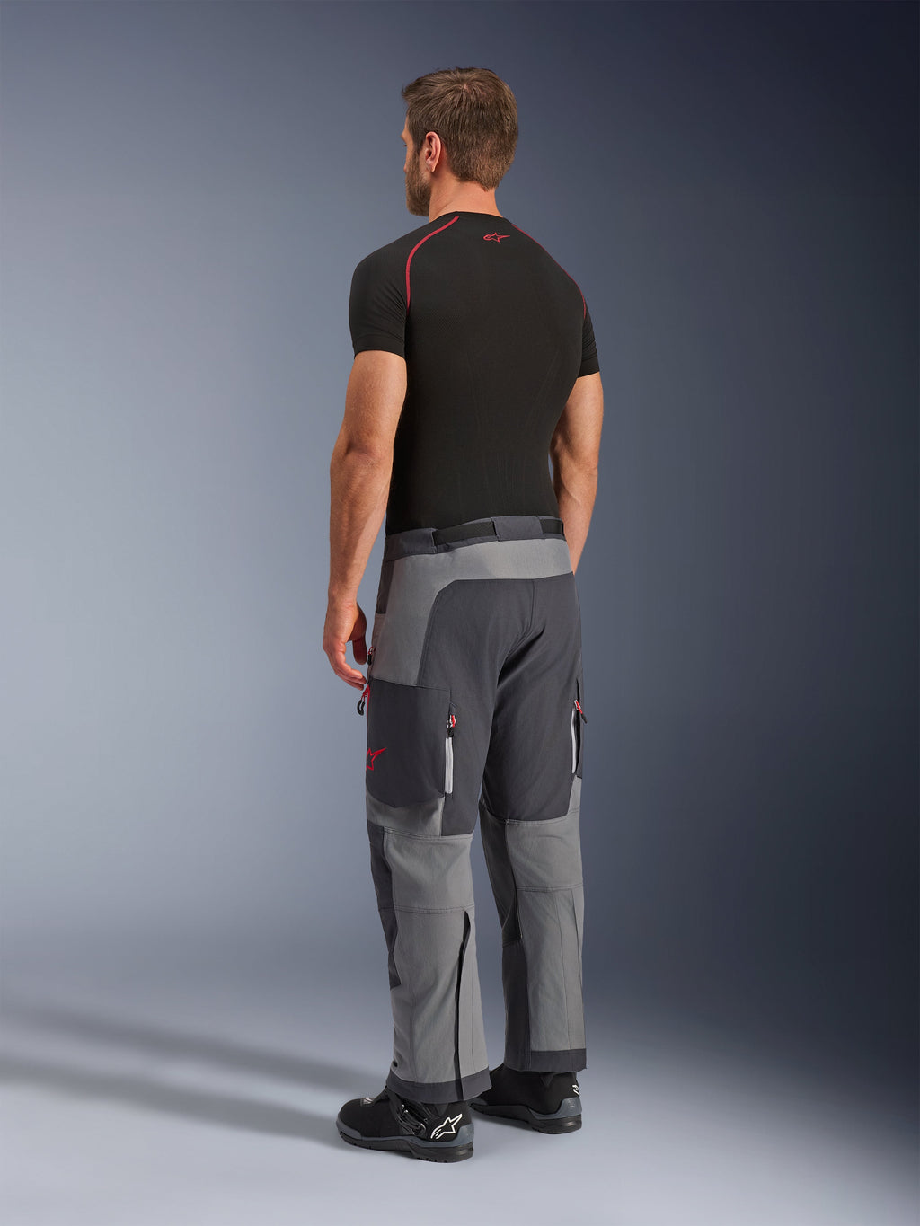 Alpinestars Maxdura Dual Pants, calças de motociclismo de aventura, cinzento escuro e cinzento profundo, vestidas por um modelo numa vista lateral traseira, com detalhes vermelhos nos fechos e no logótipo, painéis cinzentos reforçados, bolsos cargo e ajuste sobre a bota