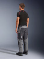 Alpinestars Maxdura Dual Pants, calças de motociclismo de aventura, cinzento escuro e cinzento profundo, vestidas por um modelo numa vista lateral traseira, com detalhes vermelhos nos fechos e no logótipo, painéis cinzentos reforçados, bolsos cargo e ajuste sobre a bota