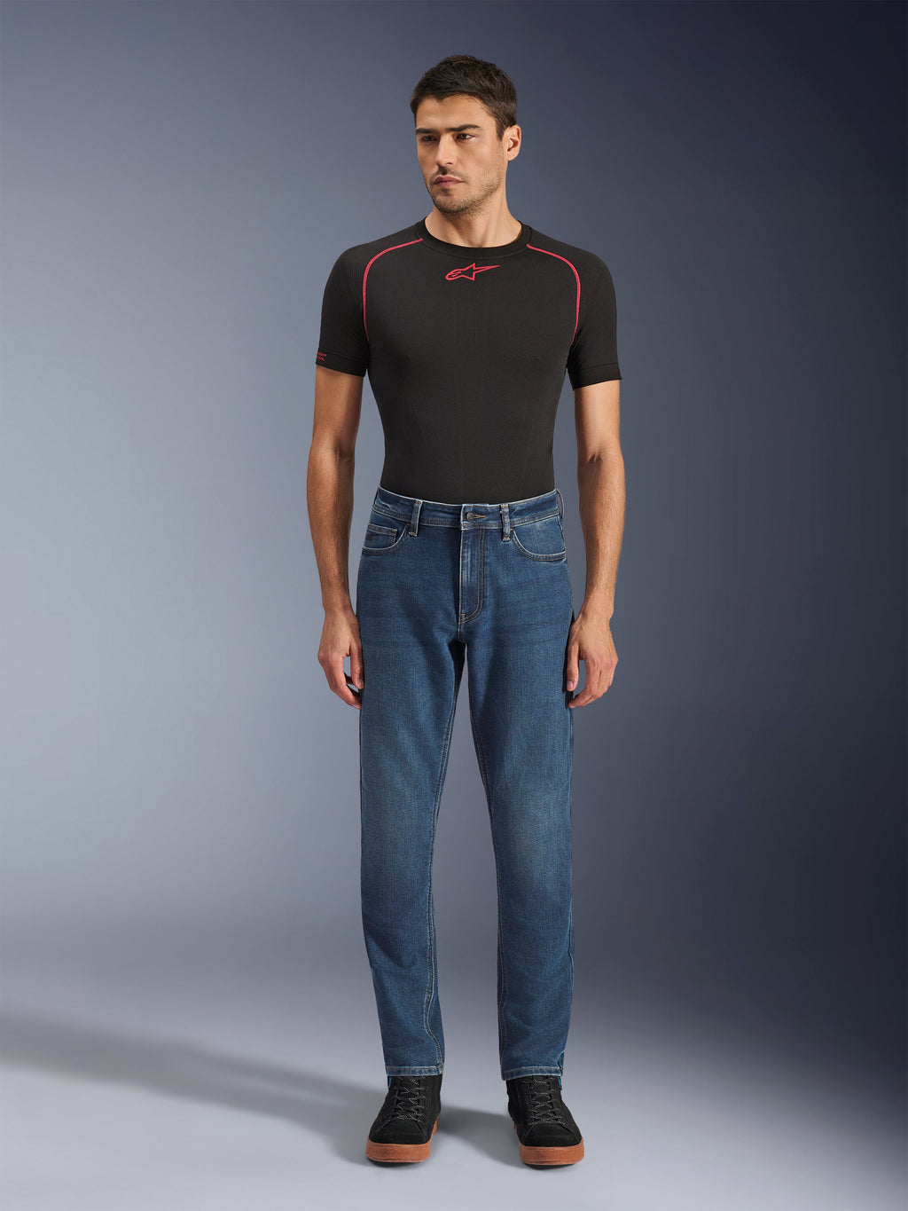 Alpinestars Flex-AST 5 Denim Pants, calças de motociclismo em ganga azul com lavagem índigo, corte regular afunilado, vestidas por um modelo com uma camada de base preta Alpinestars e sapatos casuais pretos, vestuário de estilo de vida para condução urbana.