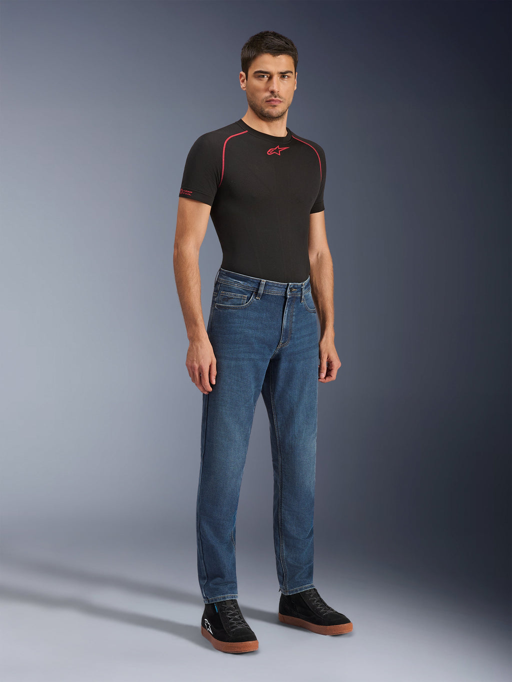 Alpinestars Flex-AST 5 Denim Pants, calças de ganga para motociclismo Indigo Washed, corte moderno afunilado, vestidas por um modelo masculino com uma t-shirt de performance preta e ténis pretos, vestuário casual de condução urbana.