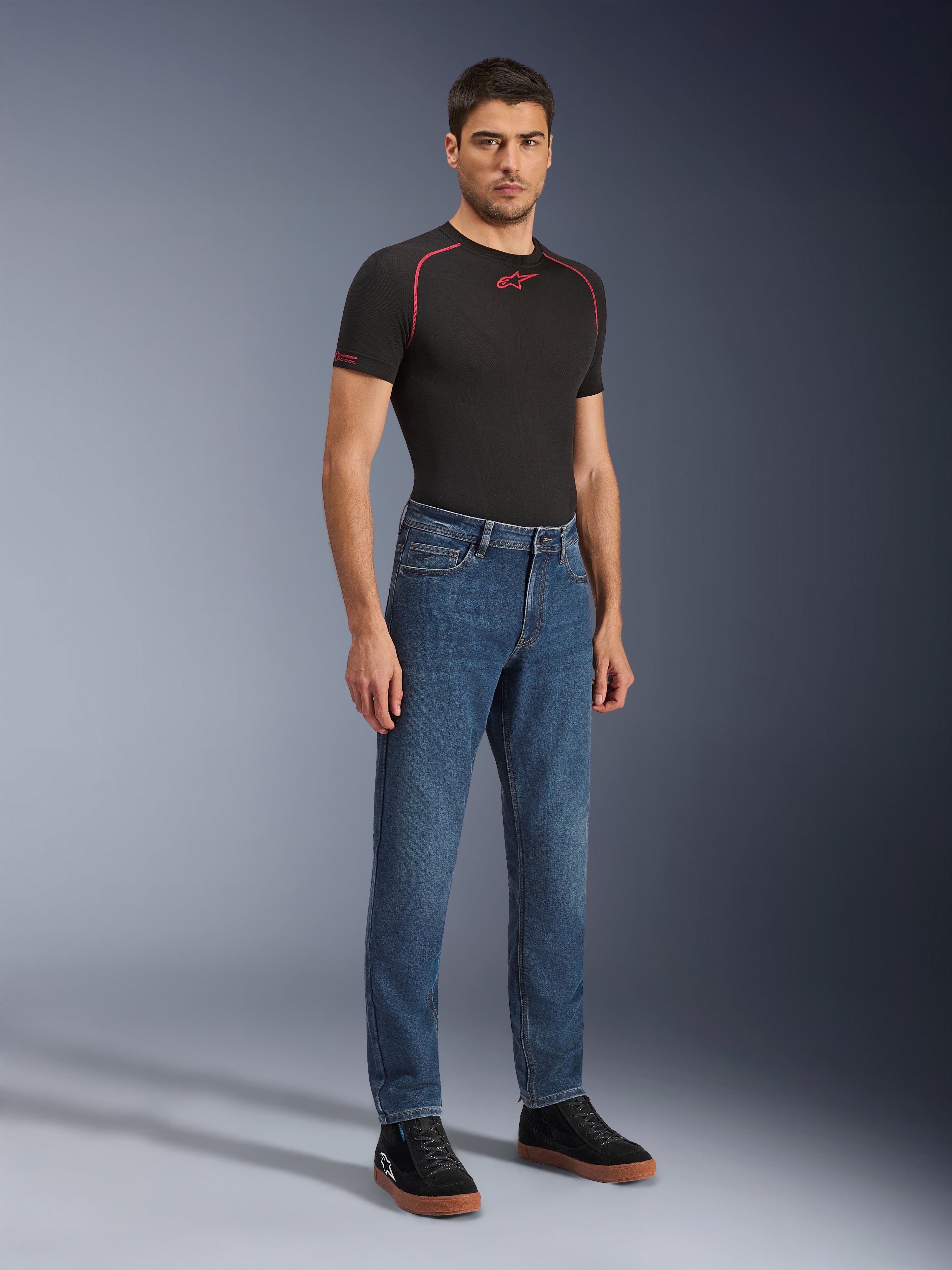 Alpinestars Flex-AST 5 Denim Pants, calças de ganga para motociclismo Indigo Washed, corte moderno afunilado, vestidas por um modelo masculino com uma t-shirt de performance preta e ténis pretos, vestuário casual de condução urbana.