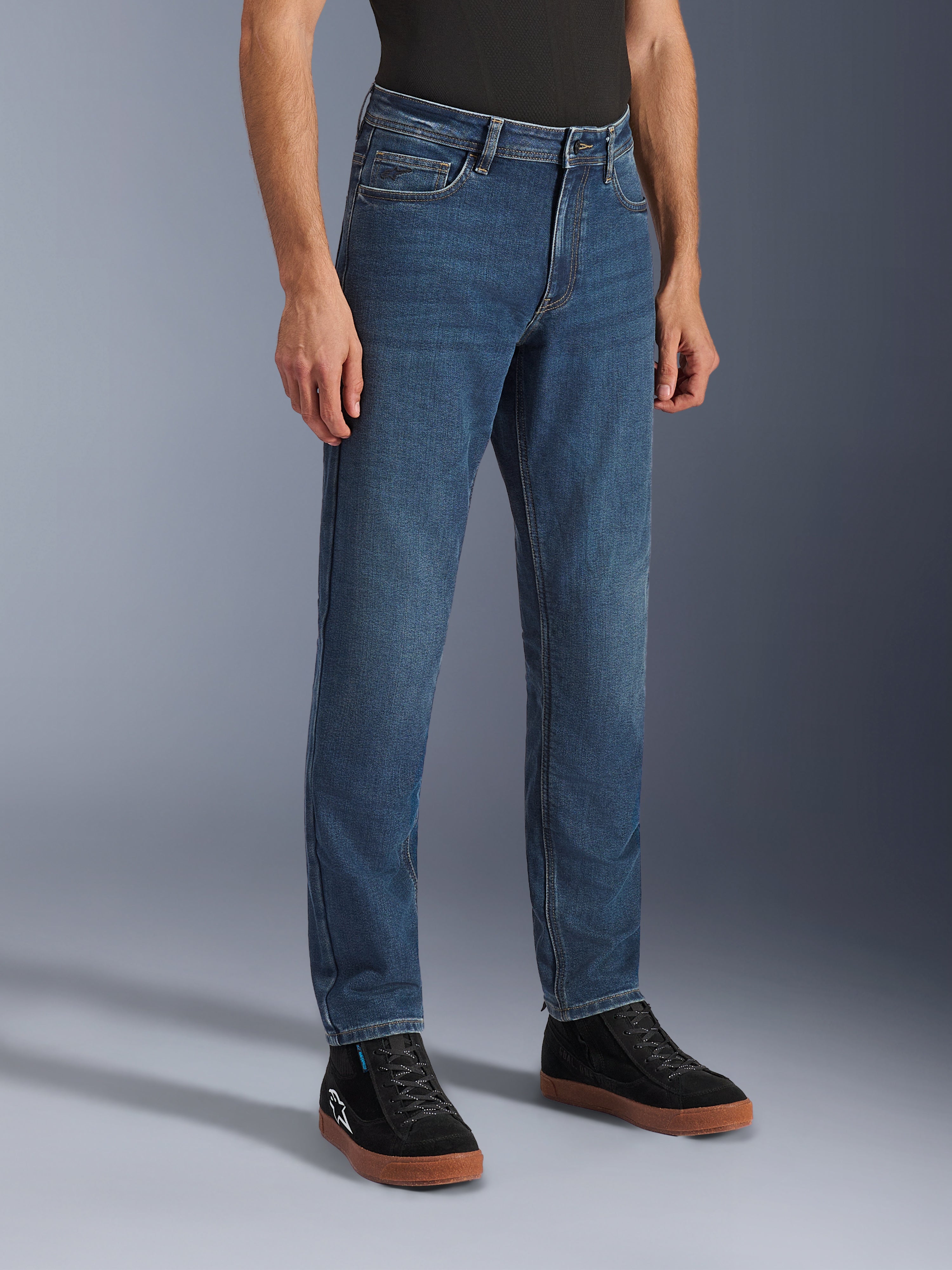 Calças Alpinestars Flex-AST 5 Denim, calças de ganga de motociclismo Indigo Washed, corte regular afunilado, usadas com sapatilhas de cano alto pretas e uma camisola preta, equipamento de condução urbana casual com um aspeto de ganga clássico.
