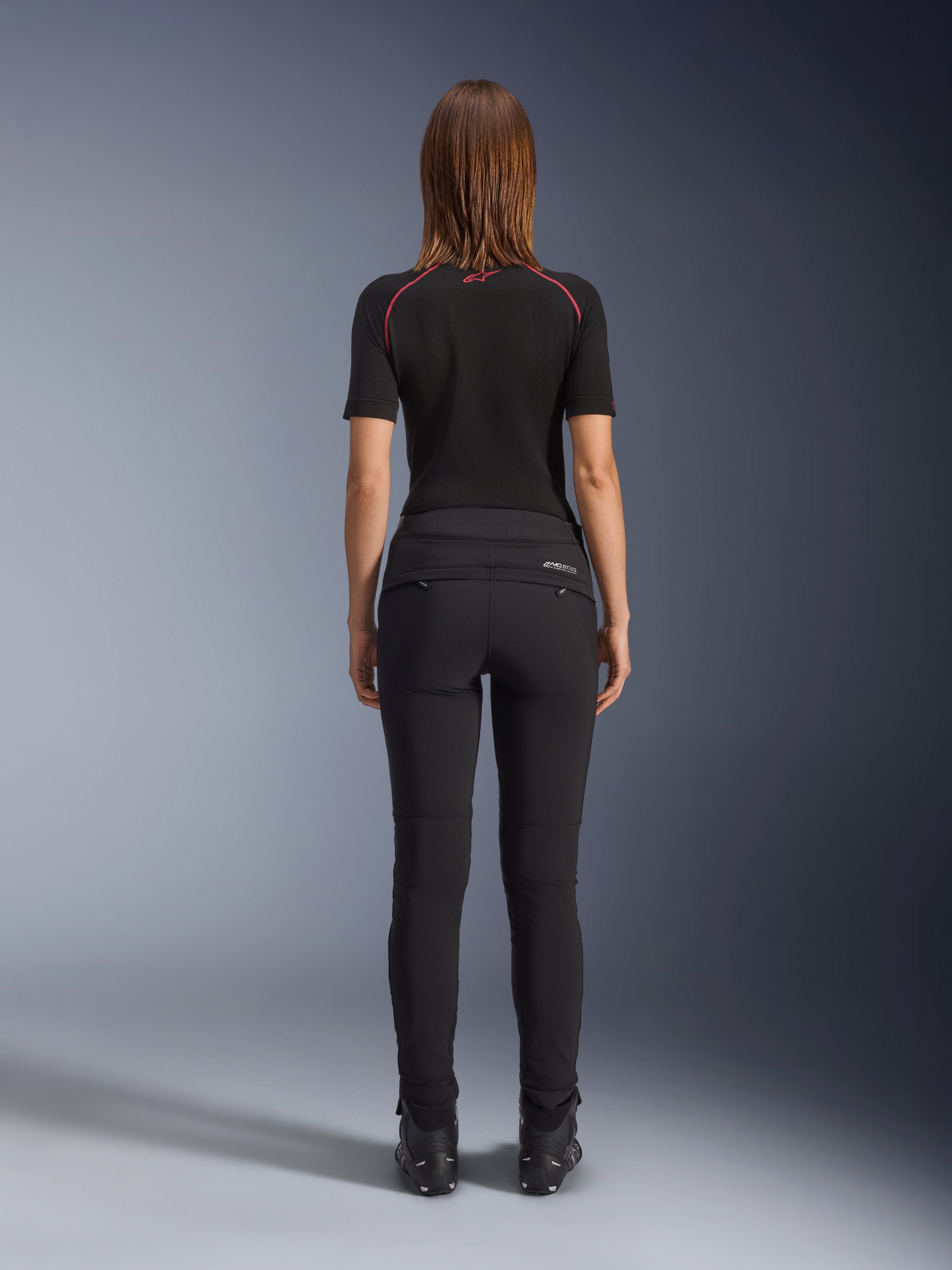 Calças Alamosa Femininas Alpinestars, pretas, vista traseira realçando a construção têxtil de corte slim, bolsos traseiros com fecho e marca refletora MO.ST.EQ. na pala traseira, usadas com uma camisola interior de manga curta preta e botas de motociclismo pretas