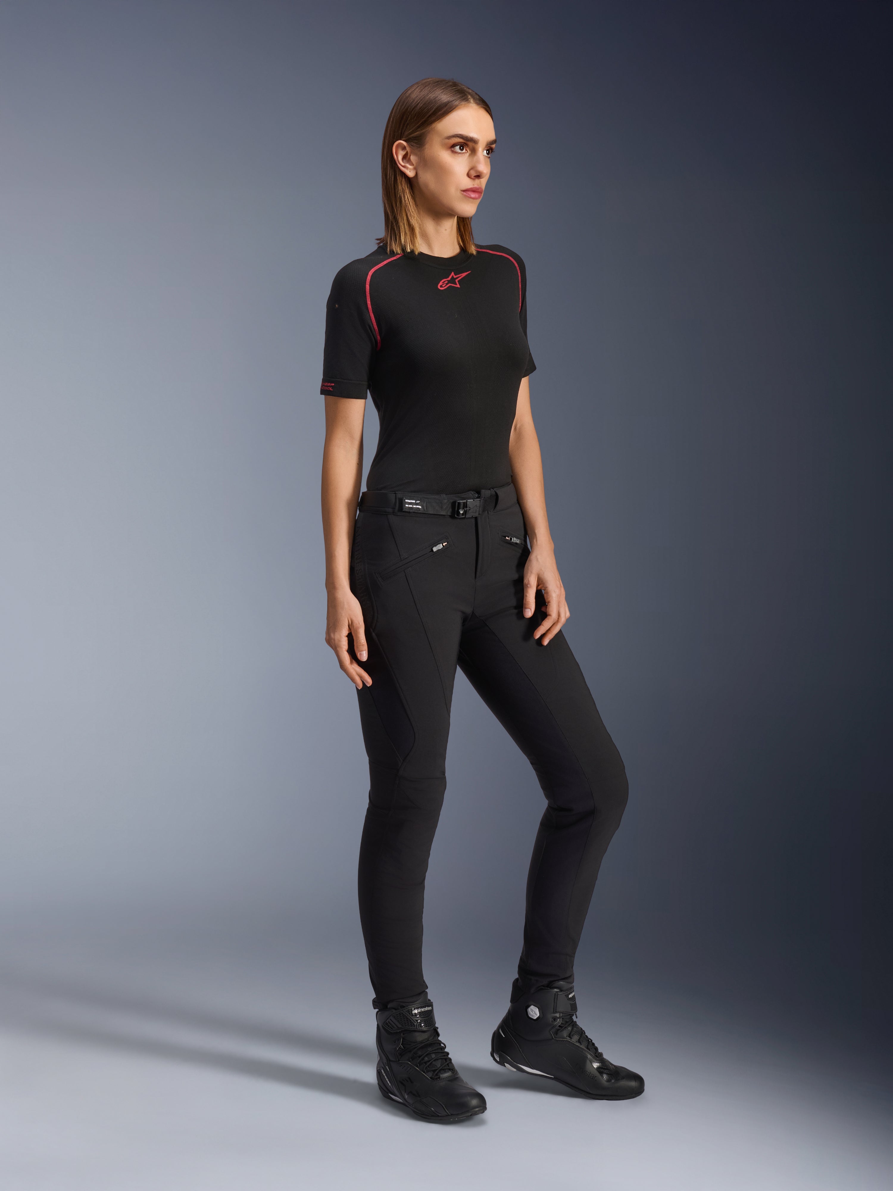 Calças Alpinestars Alamosa Femininas, calças de condução de motociclismo slim-fit em preto, vestidas por uma modelo com uma camada base preta e sapatos de condução, com bolsos frontais com fecho de correr e cinto com fecho magnético, estilo streetwear urbano.