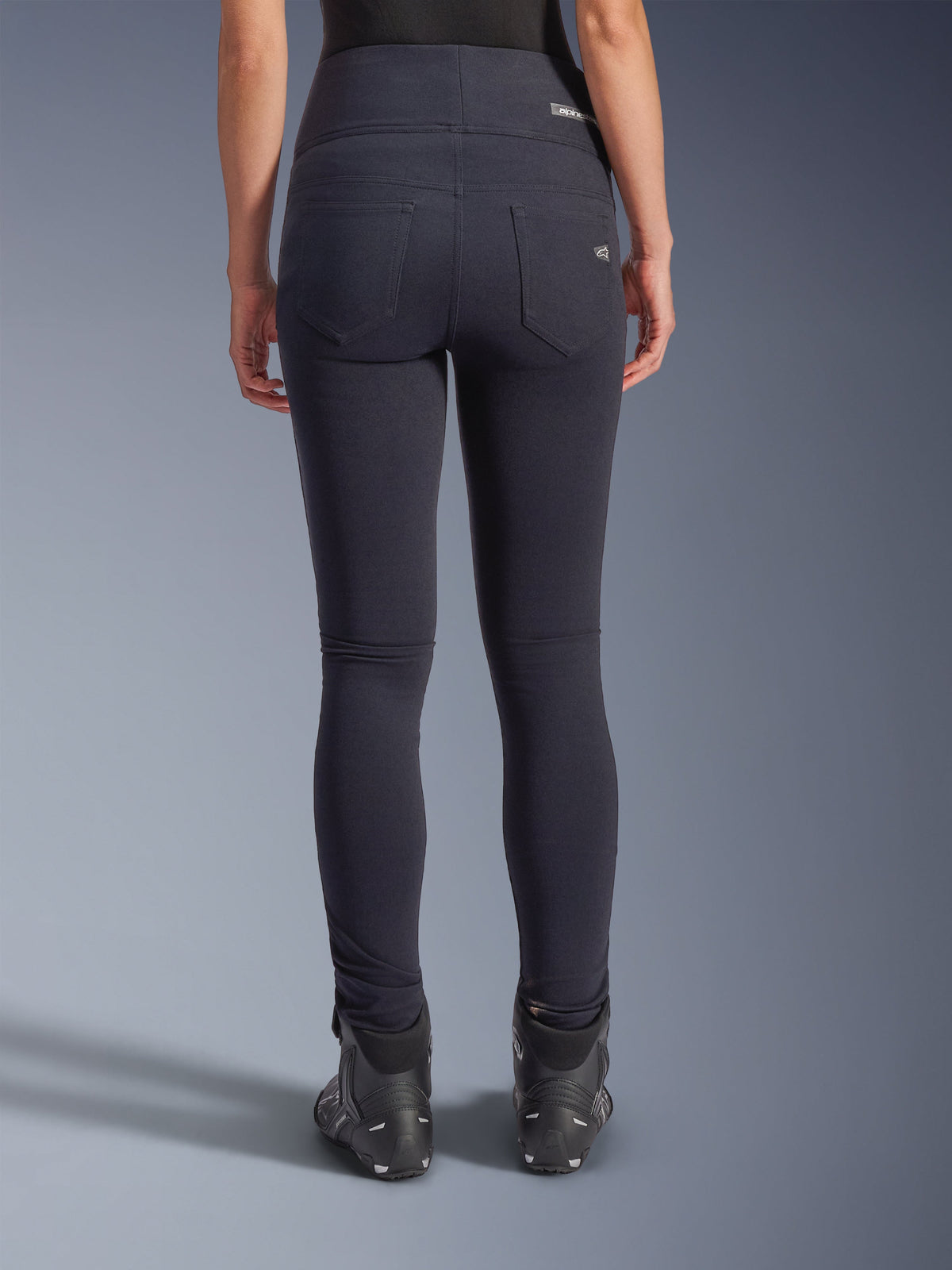 Alpinestars Stella Flex-AST Leggings, calças de motociclismo urbanas, pretas, vista traseira com corte justo de cintura alta, dois bolsos traseiros funcionais com marca subtil, material têxtil elástico, usadas com sapatos de condução pretos.