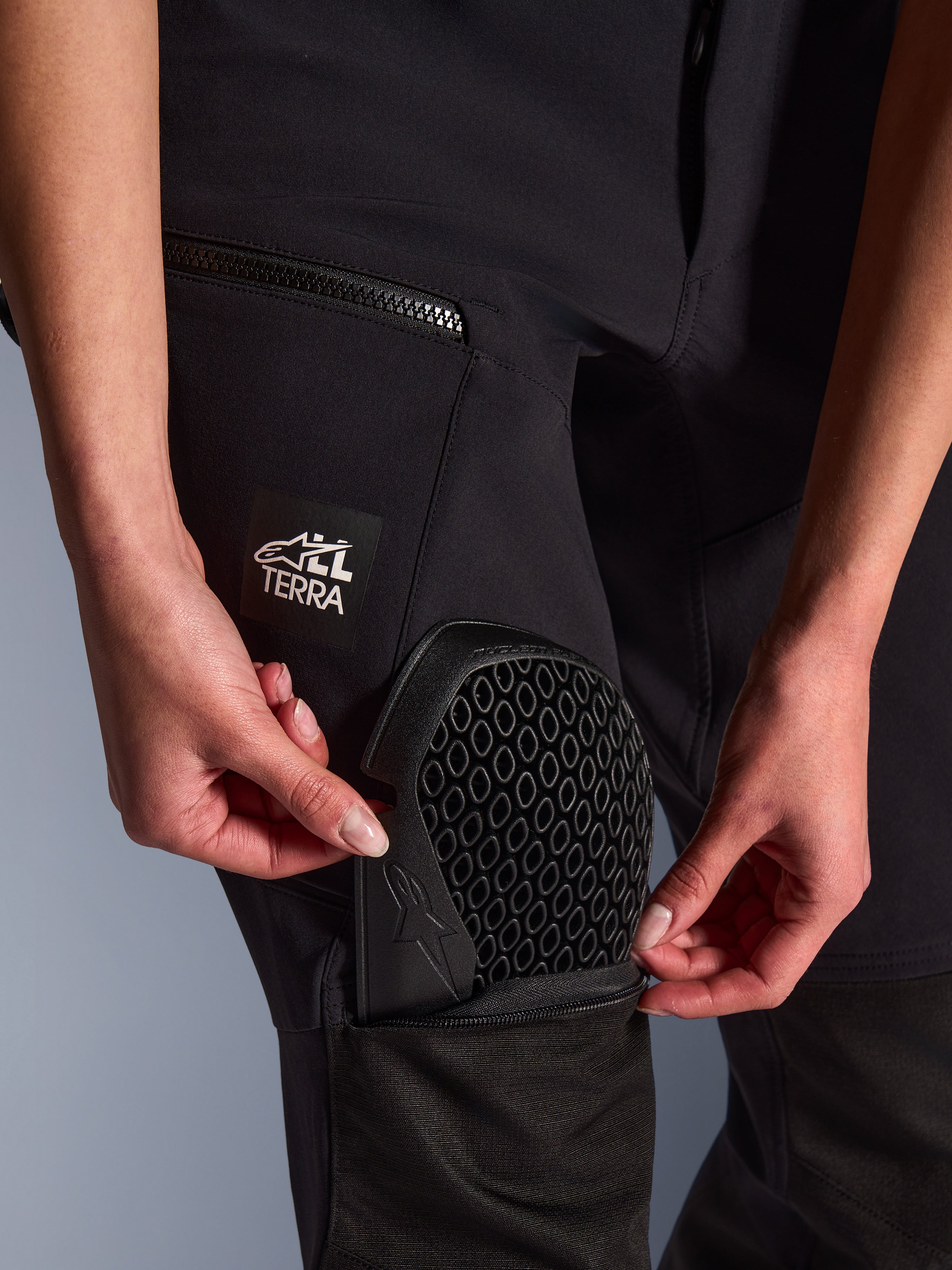 Calças Alpinestars Stella Flex-AST Explorer em preto, pormenor do bolso externo para proteções com a inserção da proteção de joelho Nucleon Flex Plus, detalhe da proteção com padrão em favo de mel, logótipo "ALL TERRA" na coxa, equipamento têxtil funcional de aventura para mulher