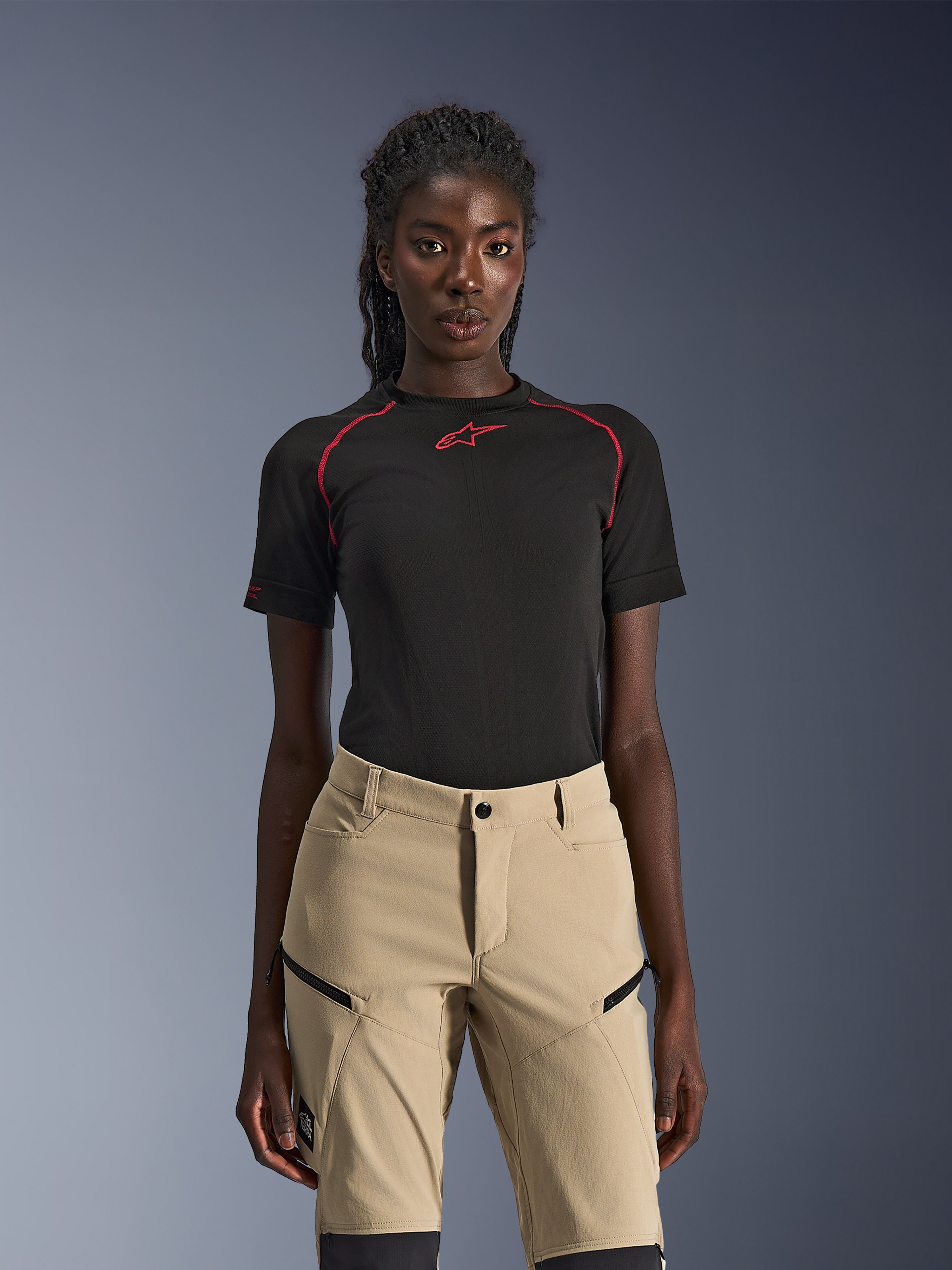 Calças Alpinestars Stella Flex-AST Explorer, calças de motociclismo para mulher em cor areia, usadas com um top técnico de manga curta preto com costuras vermelhas, equipamento de touring de aventura com bolsos cargo com fecho e tecido ripstop resistente.