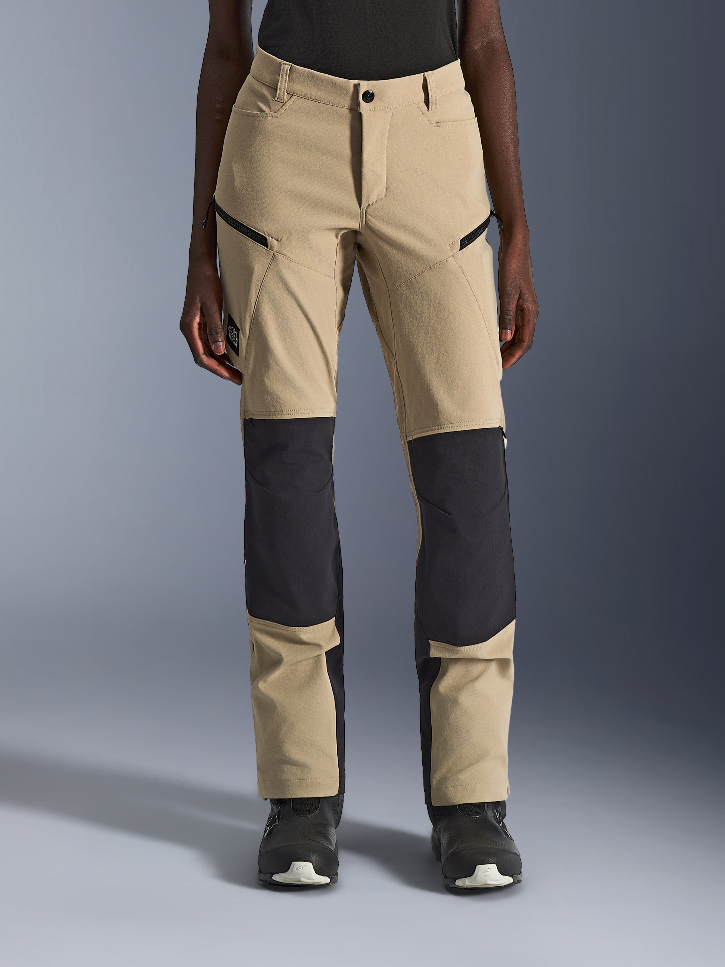 Alpinestars Stella Flex-AST Explorer Pants, calças de motociclismo para mulher, Aluminum Black, vista frontal numa modelo, com tecido bege areia e painéis ripstop pretos proeminentes nos joelhos e na parte inferior das pernas, bolsos com fecho de correr, estilo adventure touring